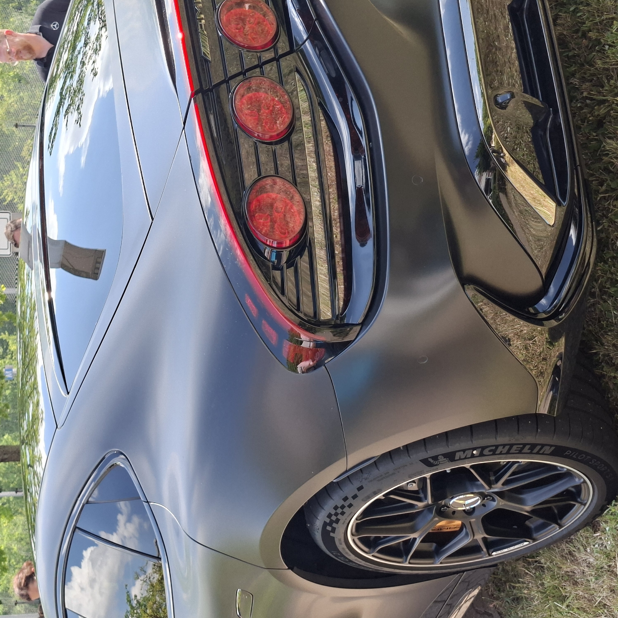 The image shows the rear quarter of a dark gray, matte finish sedan with a sleek, modern design. The car features prominent, round red taillights with intricate internal detailing, a thin red light strip across the trunk, and a black, glossy diffuser at the bottom. The car is parked on grass, with trees and foliage visible in the background. A man with glasses and a black polo shirt with a Mercedes-Benz logo is partially visible on the right, and another person's head and shoulder are visible on the far left, peeking over the car's roof. The reflection on the car's body suggests an outdoor setting, possibly a parking lot or event space. The overall scene is well-lit, indicating daytime. The question of location in Rastatt, Germany, is not directly supported by visual cues within the image itself.