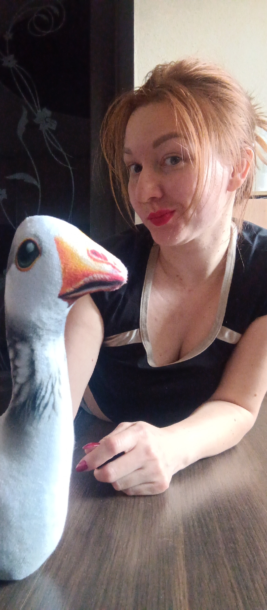 The image features a woman posing indoors with a plush goose toy. The woman is on the right side of the frame, looking towards the viewer with a gentle smile and red lipstick. She has light reddish-brown hair, styled in a way that some strands frame her face, and is wearing a black top with light-colored trim around the neckline. Her right hand, with red nail polish, rests on a dark wooden surface in the foreground.
To the left, a large plush toy resembling a goose is prominently displayed, with its head and neck facing away from the woman. The toy is light gray or white, featuring a green eye and a colorful beak that transitions from yellow to orange and red. The wooden surface in the foreground, likely a table, extends across the lower portion of the image.
In the background behind the woman, a dark wall with subtle, lighter floral or vine-like patterns is visible. To the far right, a plain lighter-colored wall appears. The lighting suggests an indoor daytime setting. There is no visible text in the image.