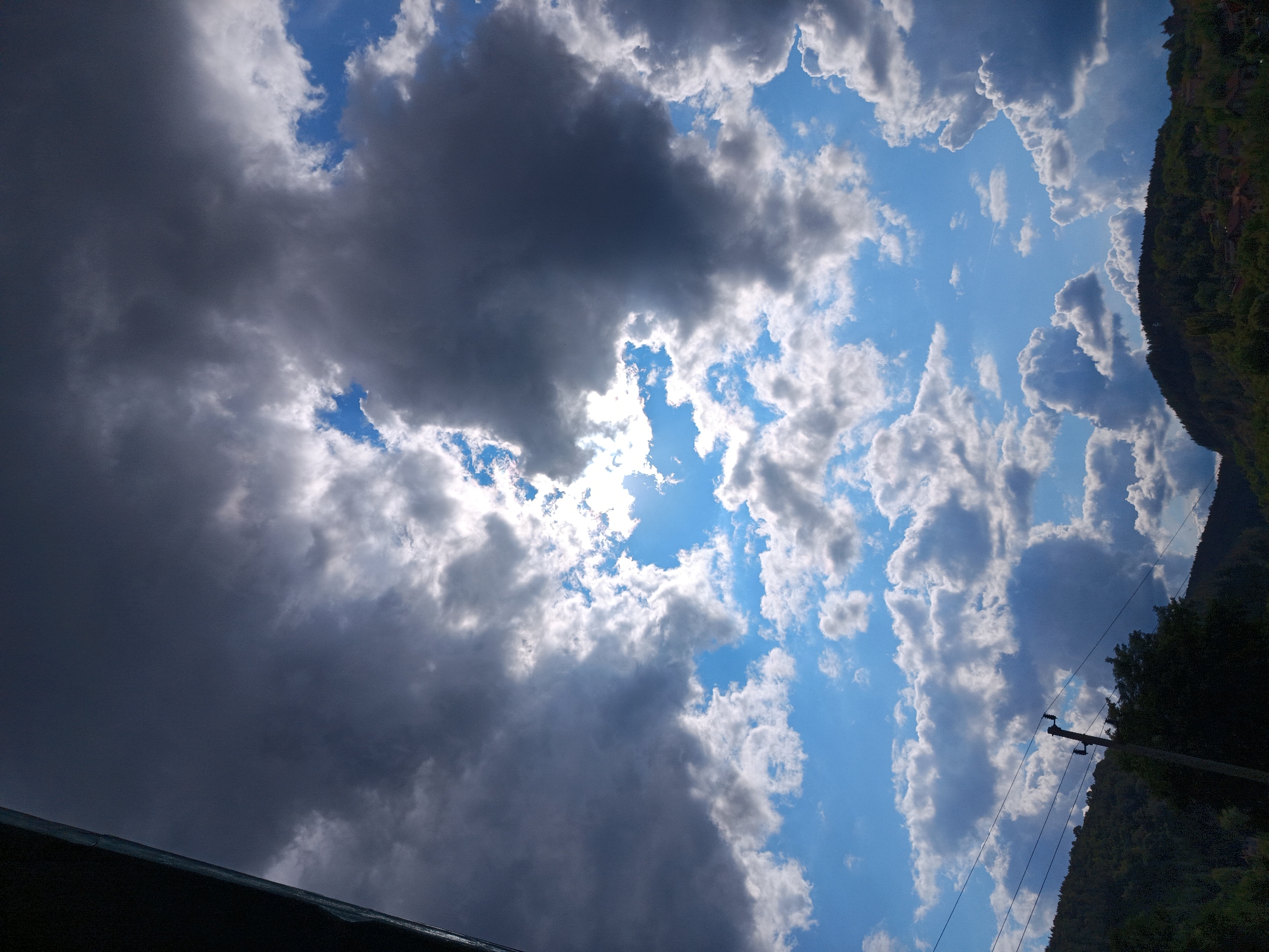 The media file displays a dramatic sky filled with a mix of dark, ominous clouds and bright, white, puffy cumulus clouds against a vivid blue sky. The sun is partially obscured by the darker clouds, creating shafts of light that illuminate the edges of the white clouds and the sky between them. To the right, a lush, green, tree-covered hillside slopes upwards, with the silhouette of trees sharply defined against the bright sky. A utility pole with visible insulators and wires runs diagonally up the hillside, suggesting a rural or semi-rural setting. The foreground is partially occupied by a dark, reflective surface, possibly a roof or a body of water, adding a sense of depth and framing the sky. The lighting and cloud formation suggest a dynamic weather system, possibly a transition from stormy to clear conditions, or vice versa. The time of day appears to be late afternoon or early evening, given the quality of the light and the angle of the sun. No people or text are visible in the image. The overall mood is one of natural grandeur and atmospheric intensity. The location context of Kralev Dol, Bulgaria, aligns with the visual cues of a hilly, forested landscape.