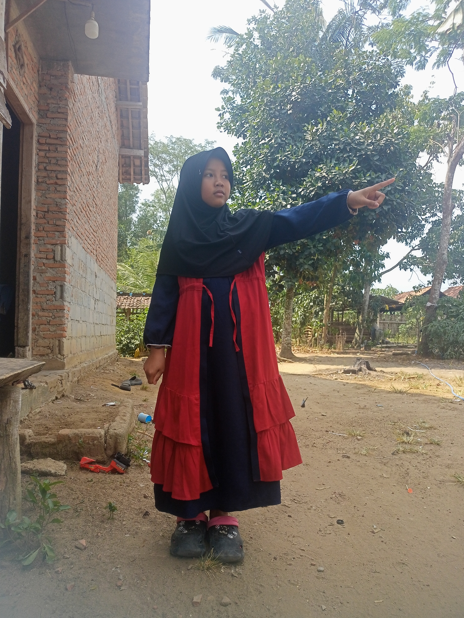 A young Indonesian girl, wearing a black hijab and a blue dress with a red overlay, stands outdoors in a dirt yard. She is pointing to the right with her right hand, her arm extended. To her left is a brick building with an open doorway. In the background, there are trees and some rudimentary structures. The ground is sandy and uneven, with scattered debris, including a red toy bulldozer and a blue plastic cup. The lighting suggests it is daytime. The scene appears to be in a rural or semi-rural area, consistent with the general environment of Pulung Kencana, Indonesia.