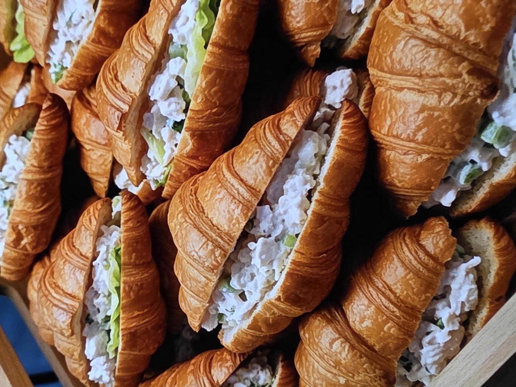 The image shows a close-up, overhead view of several chicken salad croissants. The croissants are golden brown and appear fresh, with their characteristic flaky layers visible. They are filled with a creamy chicken salad mixture containing visible pieces of chicken and possibly celery, along with some lettuce. The croissants are arranged in a close-packed display, suggesting they might be for sale. The background is dark and out of focus, drawing all attention to the food. The context of Voluntari, Romania, is not evident from the image itself; it is a detail provided externally.