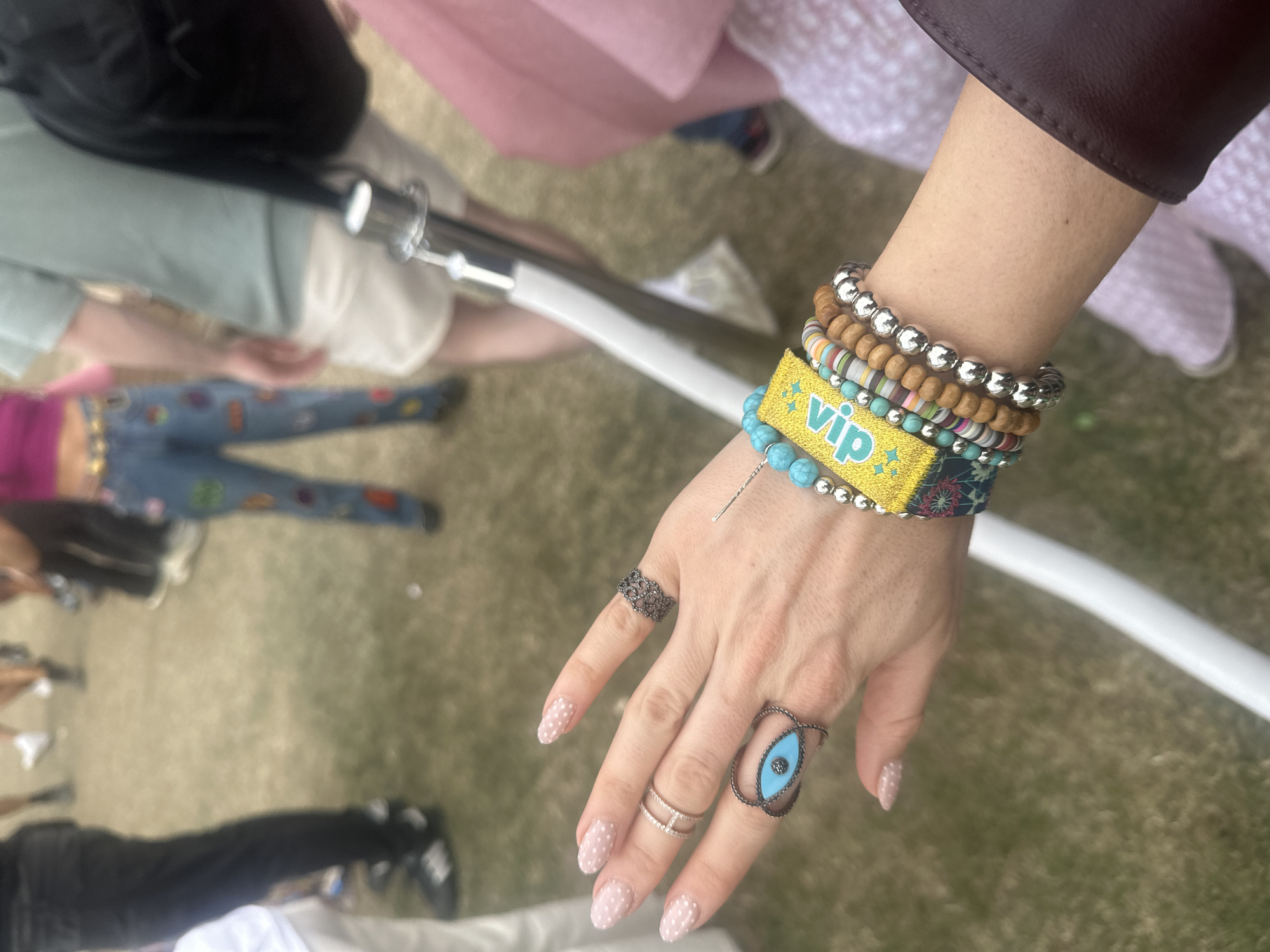 Here is a detailed description of the image:
**Scene Description:**
The image shows a close-up of a person's arm and hand adorned with multiple bracelets and rings. The person is wearing a yellow wristband with the letters "VIP" in green, along with other beaded and decorative bracelets. The hand features an oval ring with a turquoise stone on the ring finger, a double band ring on the middle finger, and a dark, ornate ring on the index finger. The fingernails are painted in a light color with white polka dots.
The person is surrounded by other people who are partially visible. There is a pair of jeans with patches on the legs, a pink skirt, and other garments. A white metal pipe is visible diagonally across the scene.
**Location:**
The exact location is unknown, and the description does not offer enough details to determine the city.