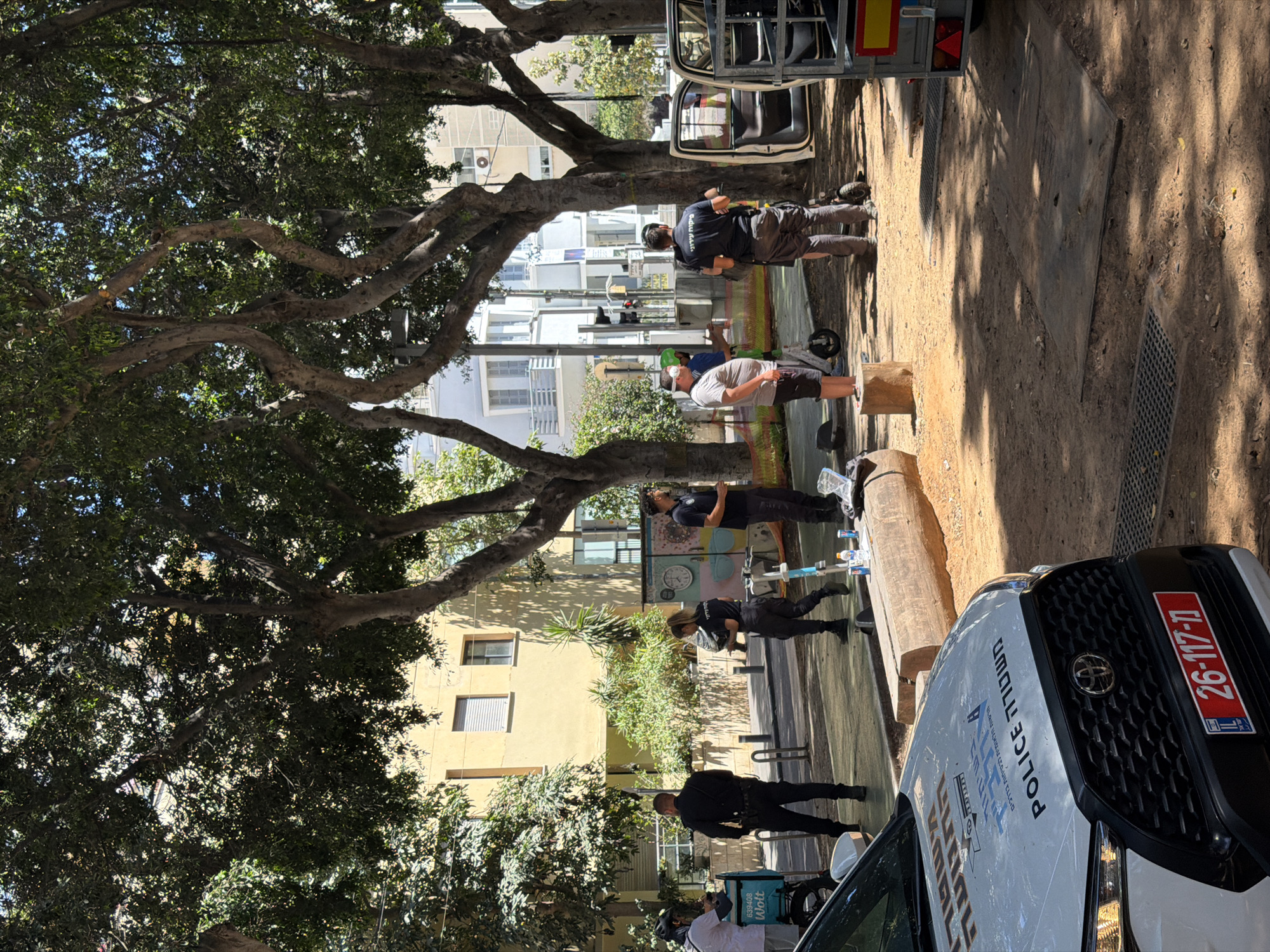In Tel Aviv, Israel, on a sunny day, a scene unfolds on a pedestrian walkway shaded by large trees. A white police car, marked with "POLICE" and Hebrew text, is parked in the foreground, its hood displaying an emblem and license plate "26-117-11". Several individuals are present. Two men in black uniforms, likely police officers, stand near the car. One man, in a light-colored shirt and shorts, is partially visible near a blue delivery bag with "Wolt" branding. Another individual in dark clothing stands near a street art mural featuring a clock. Further back, a man in a dark shirt faces away from the camera, and another man in a light-colored shirt and dark shorts stands near an electric scooter. Two other men, one in dark pants and a dark shirt with Hebrew text, and another in lighter pants, are also present. Electric scooters and various debris, including bottles and a trailer, are scattered around the area. The background consists of buildings with apartment balconies and windows.