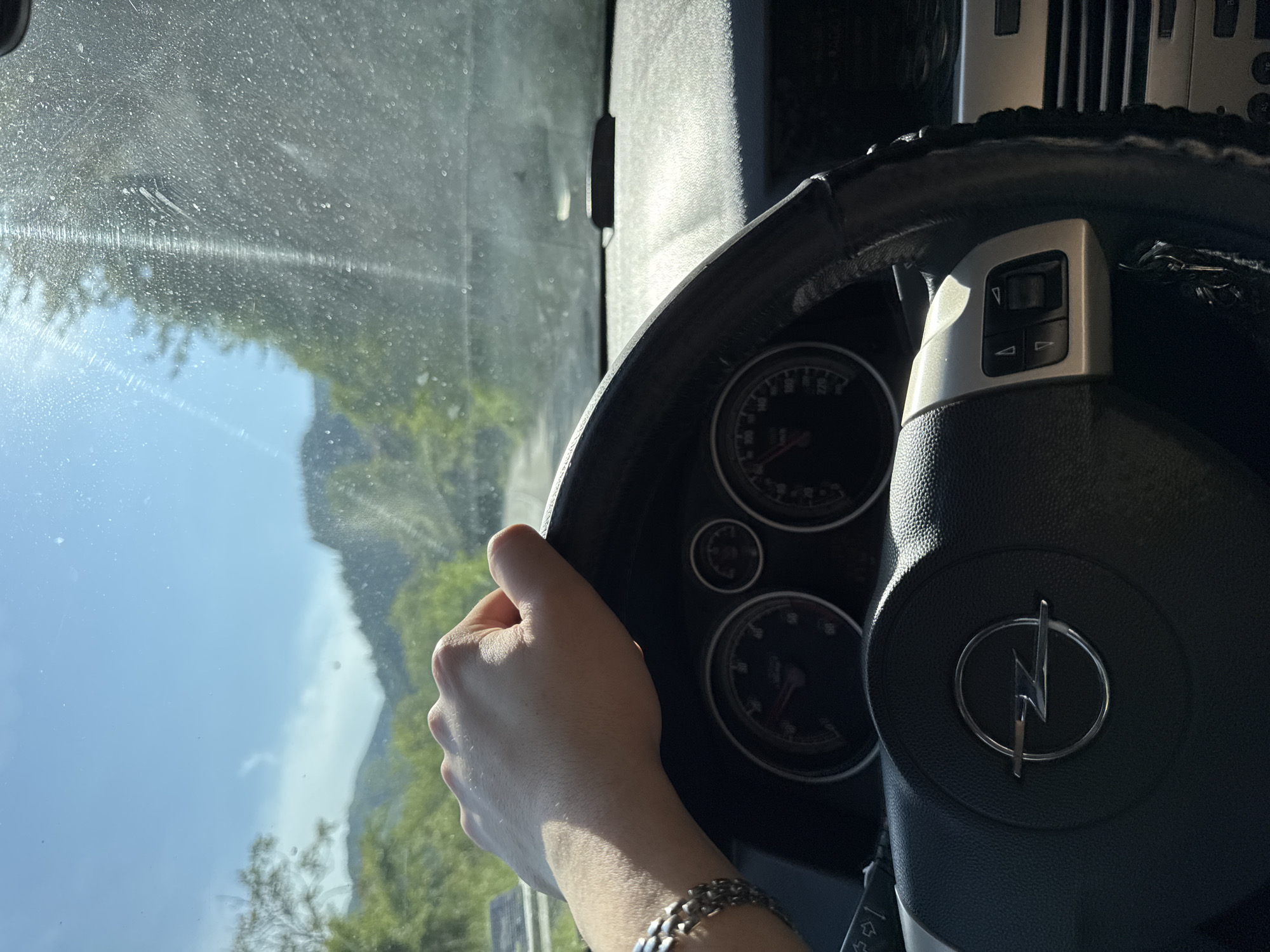 The image captures a driver's perspective from inside a car, looking out through a somewhat dirty windshield at a scenic, mountainous landscape under a clear blue sky. The scene is set in Taran, Bulgaria.
In the foreground, a human hand, presumably the driver's, is visible gripping the steering wheel. The hand is wearing a metal chain bracelet on the wrist. The steering wheel prominently features an Opel logo, characterized by a stylized lightning bolt within a circle, and has controls with arrow buttons on its left side. Part of the steering wheel is covered with a black, fuzzy material. Behind the steering wheel, portions of the car's dashboard instruments are visible, including gauges with numbers such as "100," "140," "180," "220," and "270," suggesting a speedometer and tachometer.
The windshield is noticeably dusty and streaked, with visible water marks and smudges that obscure the view slightly. Despite this, the bright daylight suggests it is daytime. Outside the car, a lush green landscape unfolds, with trees and hills or mountains rising into the distance. The sky is bright blue with scattered white clouds. The overall impression is of a journey or drive through a natural environment.