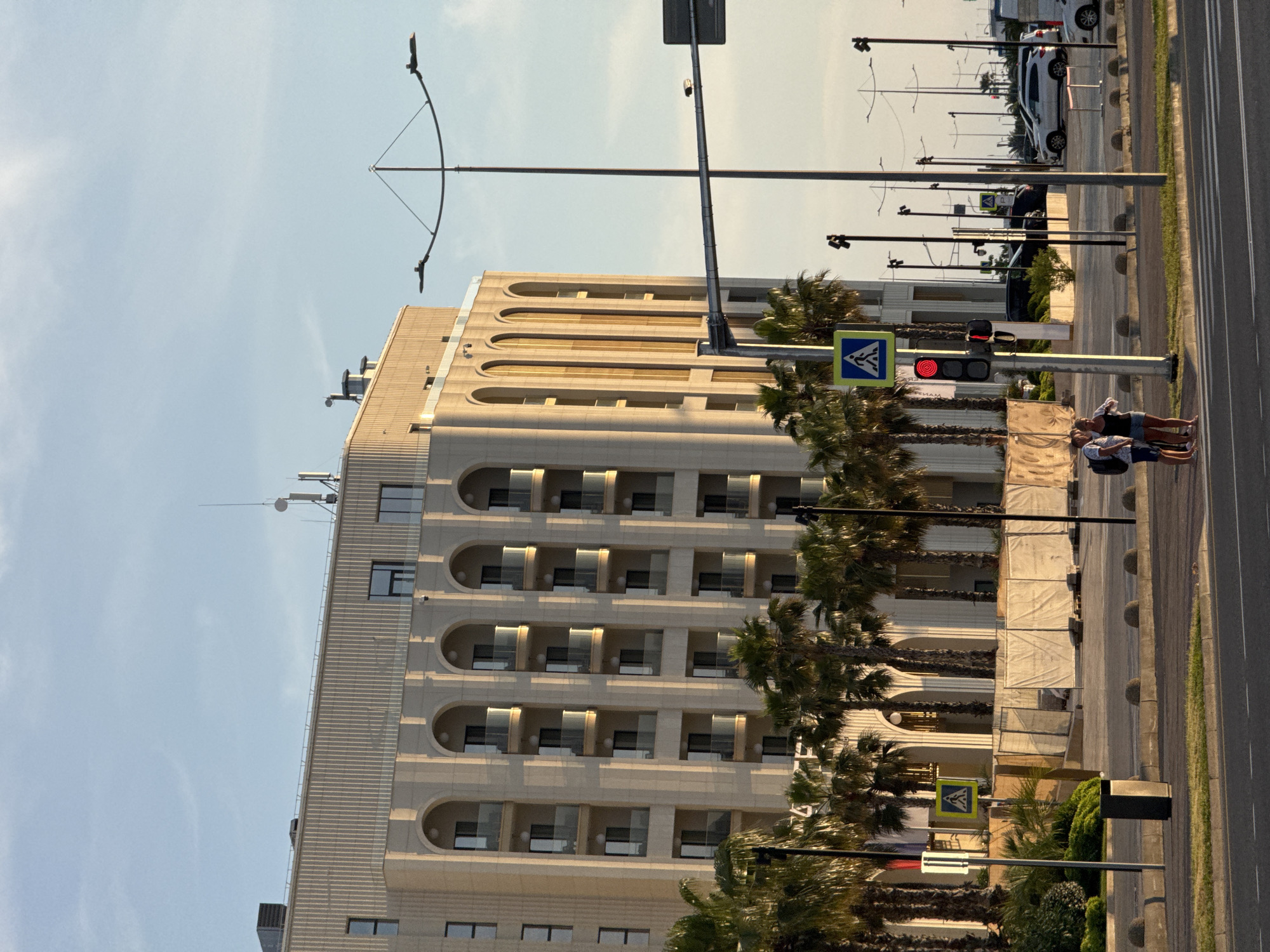 The image depicts an urban scene in Sirius, Russia, under clear sky conditions with the warm light of a late afternoon or early evening sun casting long shadows. A prominent, multi-story building with a light-colored facade dominates the background, featuring repetitive arched windows and balconies with glass railings on its lower visible levels, and a textured, ribbed upper section with rectangular windows and rooftop equipment including antennas and ventilation units.
In the foreground, a multi-lane asphalt road with white lane markings is visible, including a pedestrian zebra crossing. A red traffic light is illuminated, positioned near a pedestrian crossing sign (blue square with a white walking figure). Two individuals, a man wearing a light t-shirt and dark shorts with a backpack, and a woman in a dark top and light shorts, stand near the pedestrian crossing, engaged in conversation. They are positioned on the sidewalk, which is bordered by concrete bollards. A dark trash receptacle is also on the sidewalk.
Numerous tall palm trees line the area between the road and the main building. Behind these trees and in front of the building, a fenced-off area covered with white tarps and containing building materials suggests ongoing construction or renovation. Further back, a parking lot contains several parked vehicles, including white and dark SUVs. Overhead, a tall streetlight with a curved double-arm design extends across the road, and several shorter, dark lampposts are distributed along the roadside and within the parking area.