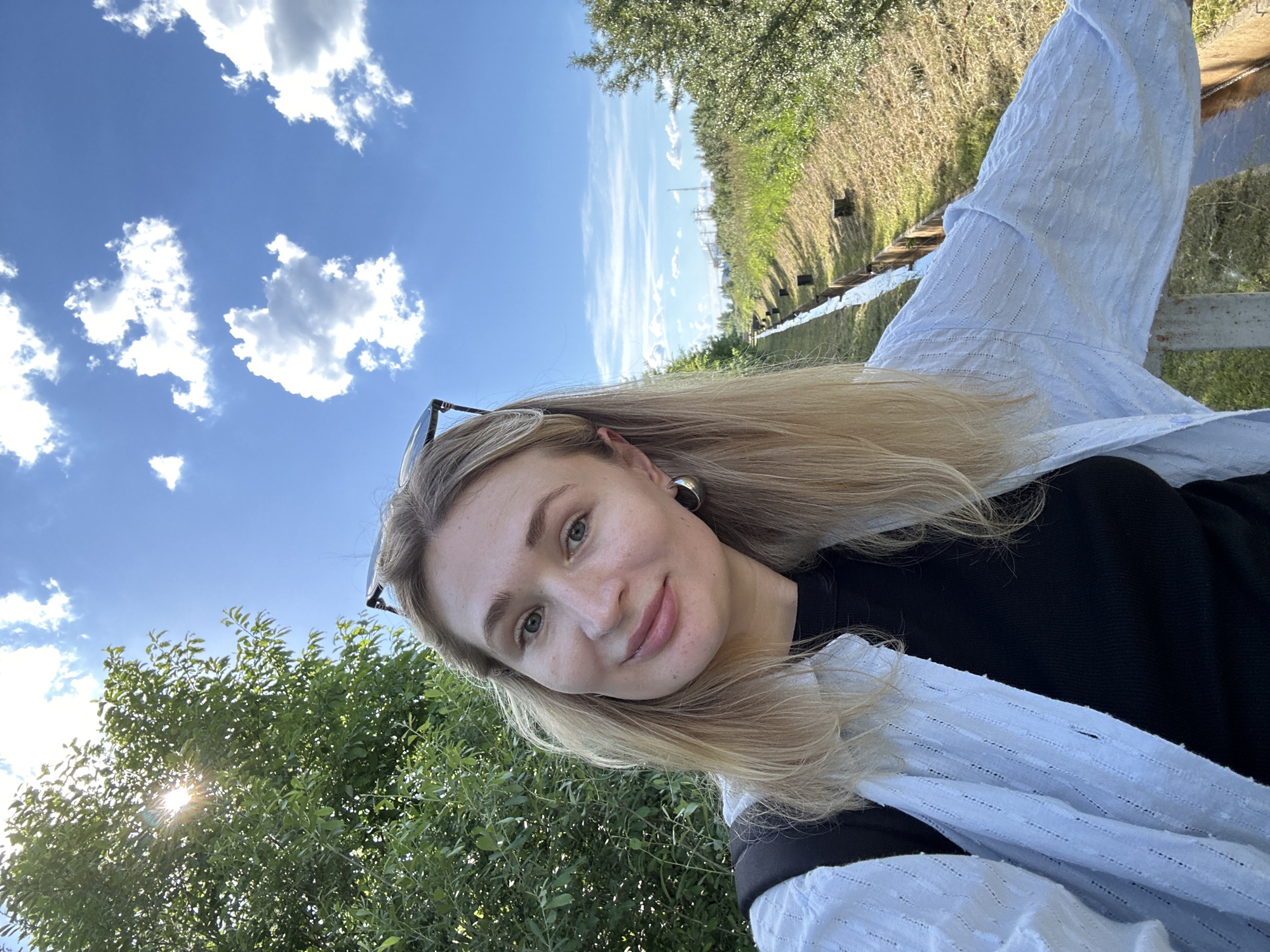 A blonde woman with fair skin and blue eyes smiles at the camera in a selfie taken outdoors on a sunny day. She is wearing a black top and a light blue button-down shirt over it. She has large hoop earrings and sunglasses perched on her head. The background features lush green trees on the left and a long, dry ditch with a small stream at the bottom on the right, bordered by a wooden fence. The sky is a bright blue with scattered white clouds. In the distance, there are power lines and more trees. The location appears to be a park or natural area within the city of Chaikovsky, Russia, possibly near the Kama River, given the prevalence of greenery and the open sky.