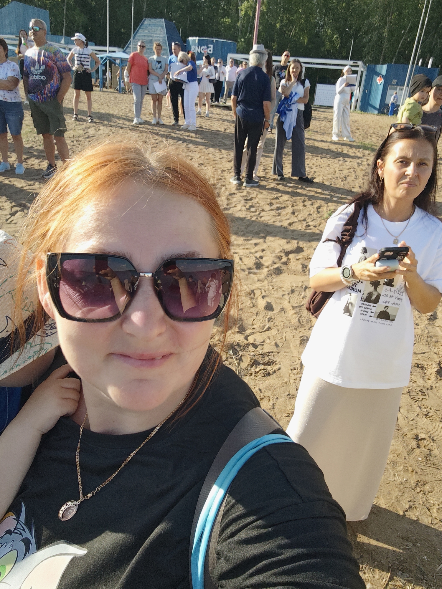 A group of people are gathered on a sandy area. In the foreground, a woman with reddish hair wearing sunglasses and a dark t-shirt with a cartoon character is visible. Behind her, to the right, another woman with dark hair, wearing a white t-shirt and a light-colored skirt, holds a mobile phone. Other individuals are seen in the background, some standing and conversing, with structures and trees in the distance. The environment appears to be outdoors, possibly a park or public gathering space, with sandy ground and a backdrop of greenery and some industrial-looking structures. The image captures a moment of casual interaction among a group of people in an open, outdoor setting.