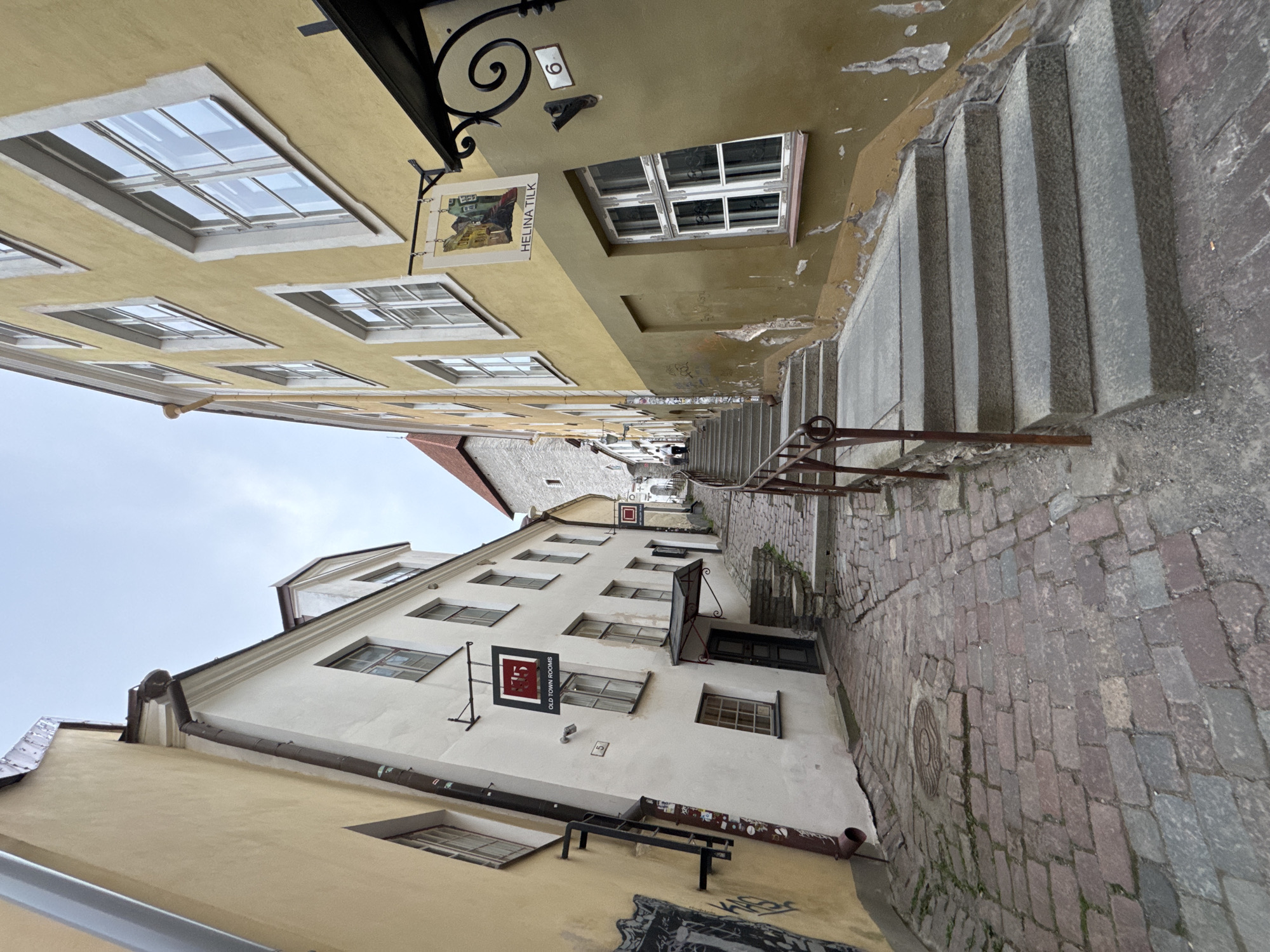 The image shows a narrow cobblestone street in Tallinn, Estonia, with historic buildings on either side. The street ascends a gentle slope, with a set of stone stairs in the foreground to the right, leading upwards. The buildings feature aged facades with visible signs of wear and tear. On the left, a cream-colored building has a dark pipe running down its side and graffiti near the bottom. A sign reading "OLD TOWN ROOMS" with the number "15" is visible. On the right, a yellowish building has a sign with a painting and the name "HELINA TILK" written below it. Above the door of the building on the right, a decorative metal awning is present, and the number "6" is displayed on a white plaque. The sky is overcast, suggesting a cloudy day. A few people are visible further up the street, walking away from the camera. The overall scene conveys a sense of historical charm and a typical urban landscape of an old European city.
