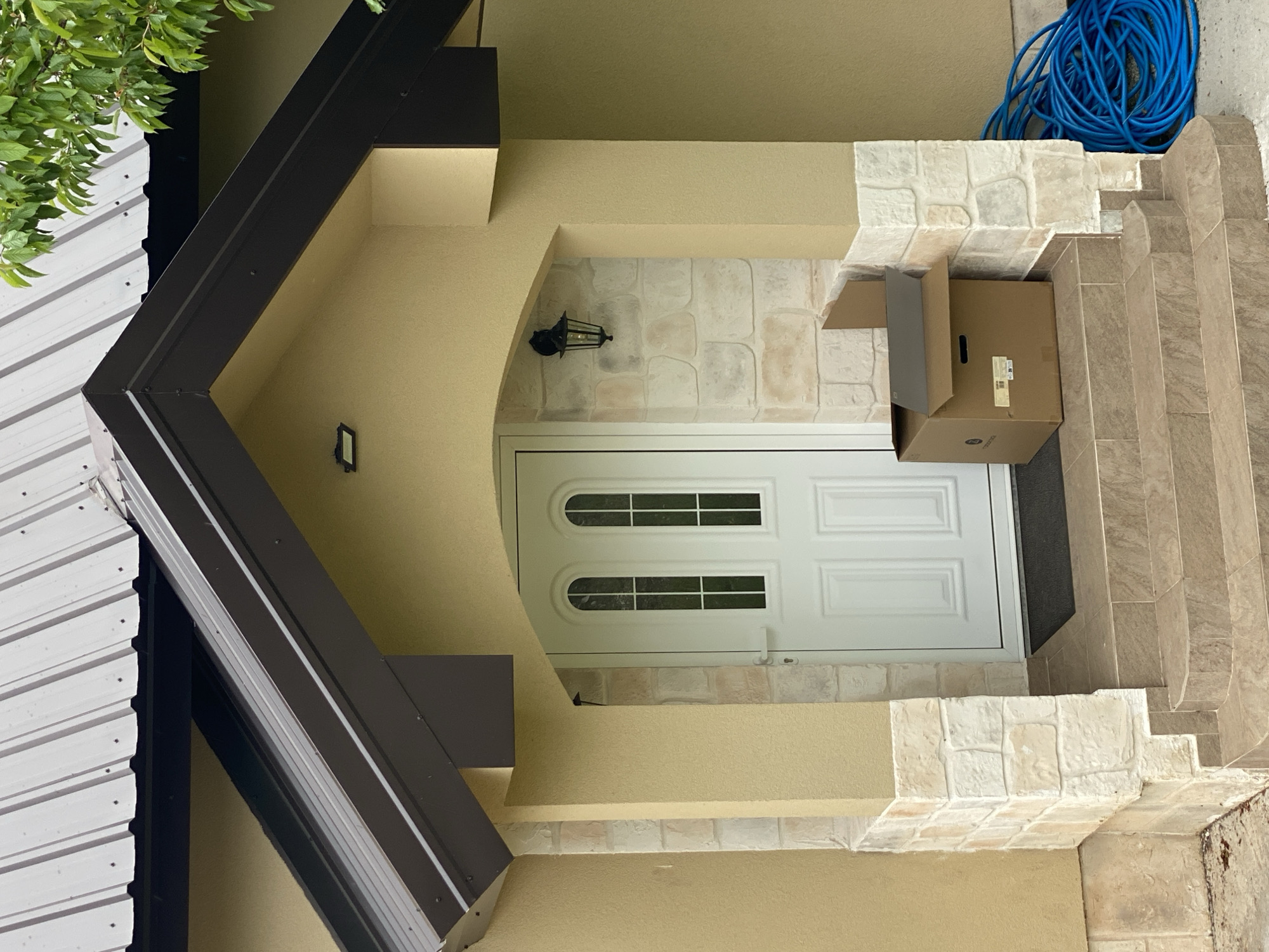 An eye-level shot captures the entrance to a building, specifically a front door accessed via a set of tiled steps and a small porch. The scene is dominated by a white, paneled front door featuring two horizontal rows of rectangular glass inserts. The door is set within a light beige, textured wall that forms an arched alcove.
To the right of the door, a series of light brown tiled steps ascend, culminating in a small landing where a coiled blue hose rests. A cardboard box, partially open and apparently empty, sits on the steps immediately to the right of the door. Visible text on a label on the box includes a barcode and the word "UPS". The box also has a small, dark logo that is difficult to decipher.
Above the door, a black, ornate lantern-style light fixture is mounted on the wall, which is adorned with light-colored, faux-stone tiles. A smaller, simpler rectangular light fixture is also visible on the left wall of the porch. Dark brown, angular eaves or soffits frame the upper left and bottom left portions of the image, suggesting an overhang or roof structure. To the far left, green foliage from a tree is visible, partially obscuring a white corrugated metal wall or roof. The ground and the steps appear to be dry, suggesting fair weather. The overall impression is a quiet, functional entrance in Korita, Montenegro, likely captured during daytime.