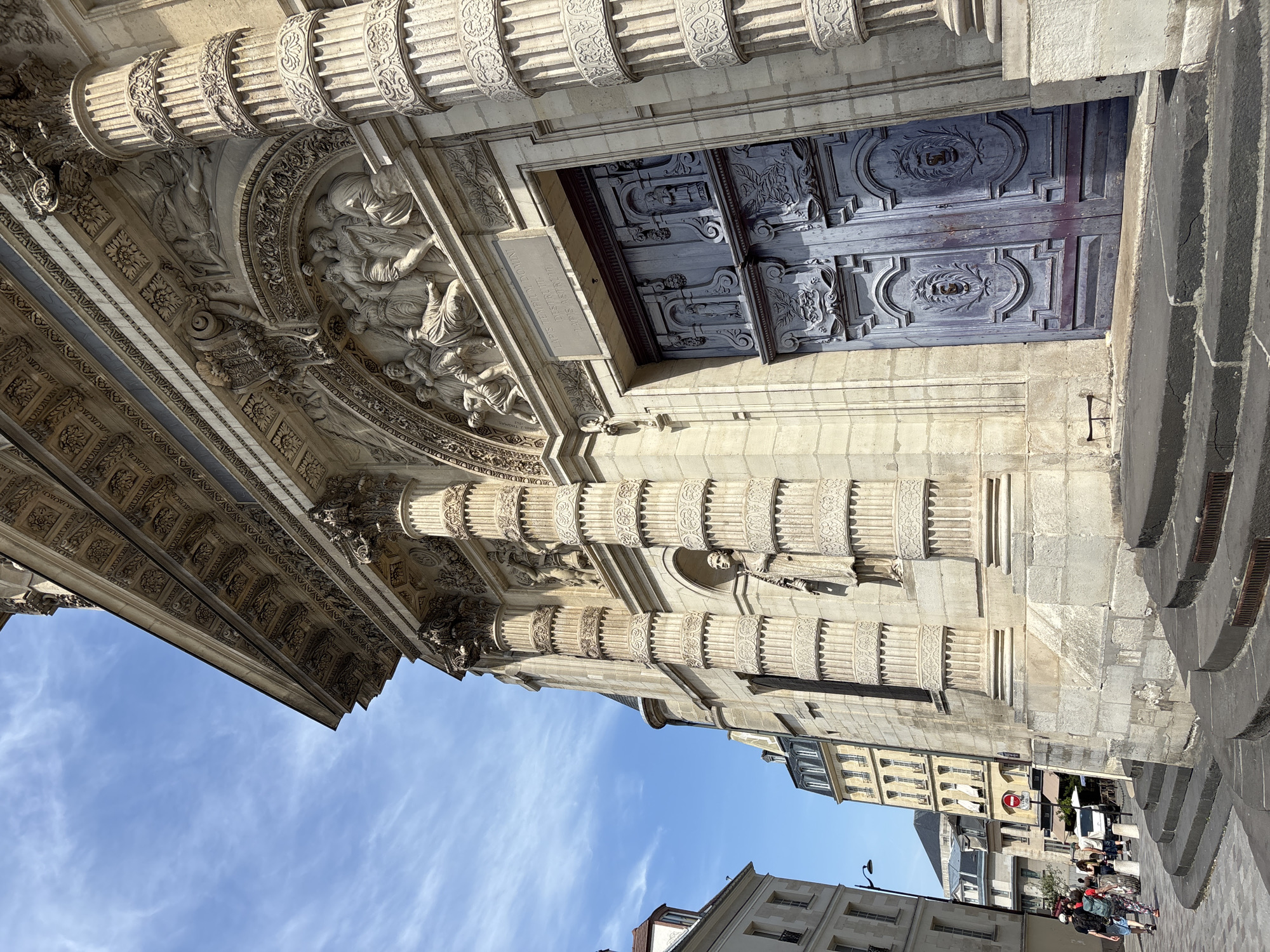 The image shows the façade of the Saint-Eustache Church in Paris, France, on a clear, blue day with wispy clouds. The church's exterior features ornate stone carvings and large, detailed double doors painted a faded purple. The doors are set within a grand entrance flanked by massive, fluted columns. To the left of the church, a street scene unfolds with people walking and a line of buildings in the background, some with mansard roofs, characteristic of Parisian architecture. The foreground shows stone steps leading up to the church entrance. The overall impression is of a historic and impressive architectural landmark.