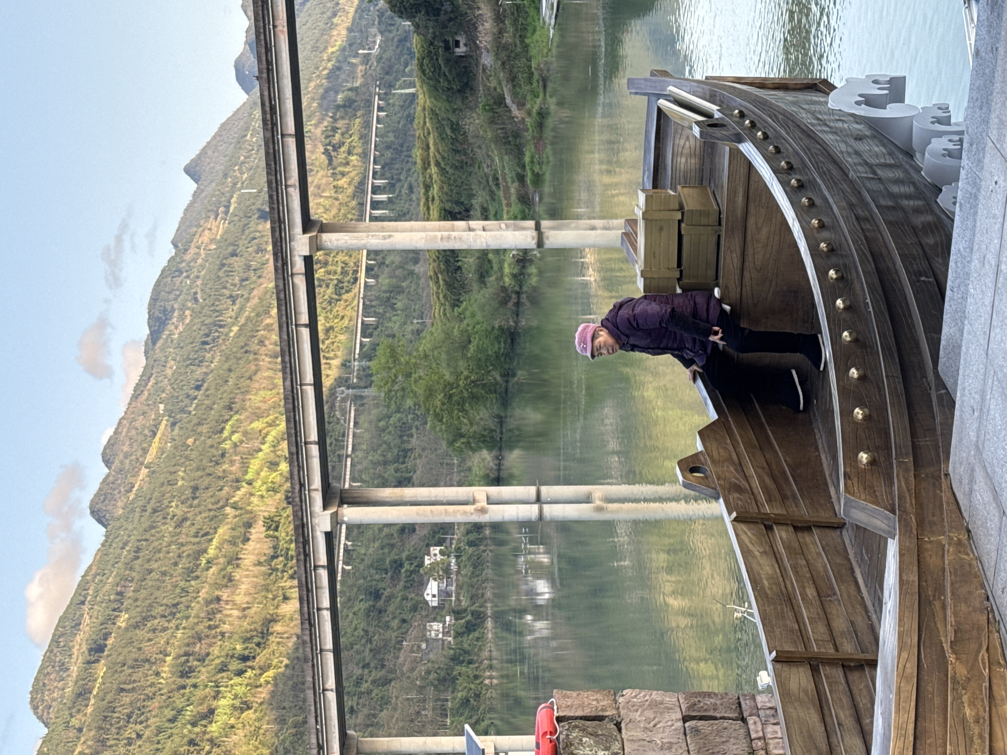 A woman wearing a purple jacket and pink hat leans forward inside a wooden boat structure next to a concrete embankment, with a body of water behind her and a bridge spanning the water in the city of Zhuoshui, China. Mountains rise behind the bridge and river, visible against a clear sky. The boat is constructed of dark wood planks and features decorative elements.