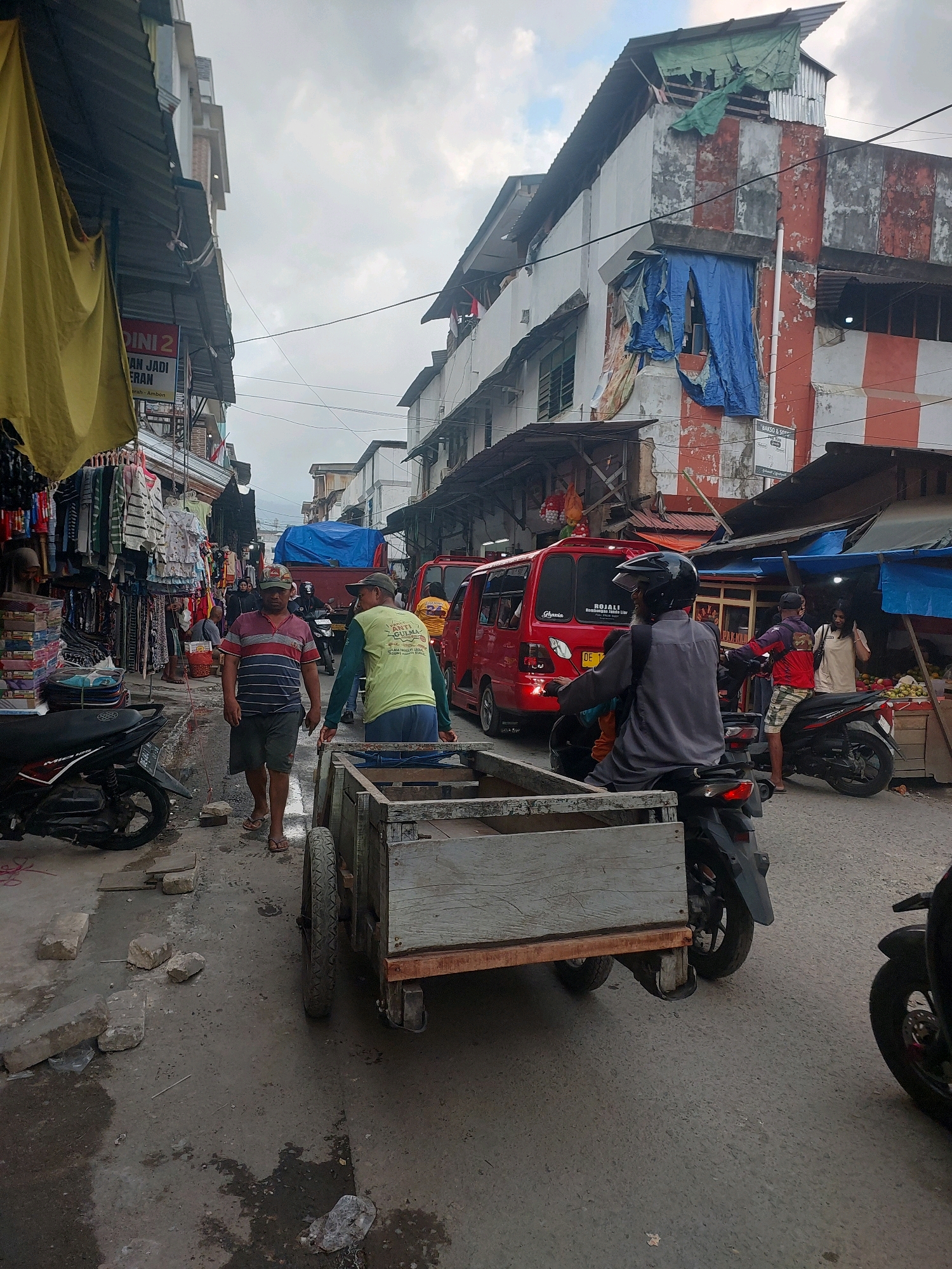 The image depicts a street scene in the city of Ambon, Indonesia. A narrow, paved street is lined with buildings on both sides. The buildings appear to be a mix of shops and residential structures, with some showing signs of disrepair and makeshift repairs using tarpaulins and corrugated metal. The sky is overcast.
In the foreground, a man in a striped shirt and shorts is walking on the street, followed by a man in a light green shirt. They are near a wooden cart with two large wheels. To the left of the cart, a dark-colored motorcycle is parked. Further down the street, a red van is visible, with another red vehicle behind it.
On the right side of the street, there are market stalls with goods displayed, including fruits. Several people are present in the scene, including individuals on motorcycles and on foot. One person on a motorcycle, wearing a helmet and a grey long-sleeved shirt, is facing away from the camera. Another person on a motorcycle, wearing a red shirt and shorts, is also visible.
The overall impression is a bustling urban environment, likely a market area, with everyday activities taking place. The presence of the cart suggests transportation of goods, common in such settings. The architecture and the surrounding environment are indicative of a tropical climate.