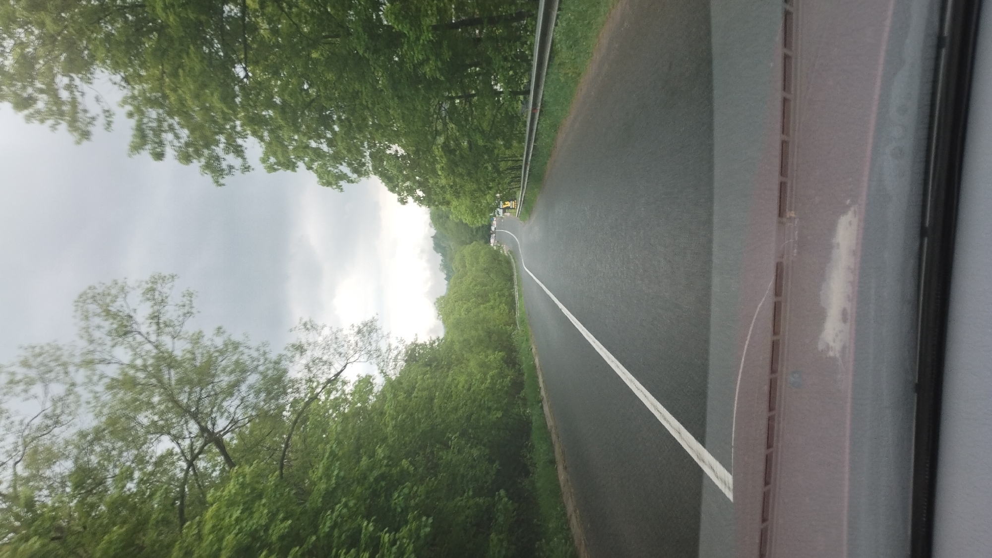 The image depicts a view from inside a vehicle driving on a road bordered by lush green trees. The road has a white line marking its center and curves slightly to the right. The sky is overcast with gray clouds. In the distance, several vehicles are visible on the road, and there are some signs and railings along the side. The foreground shows the dashboard of the vehicle. This scene is consistent with the natural surroundings found in and around Lazeshchyna, Ukraine, a village located in the Carpathian Mountains known for its scenic beauty and forested landscapes.