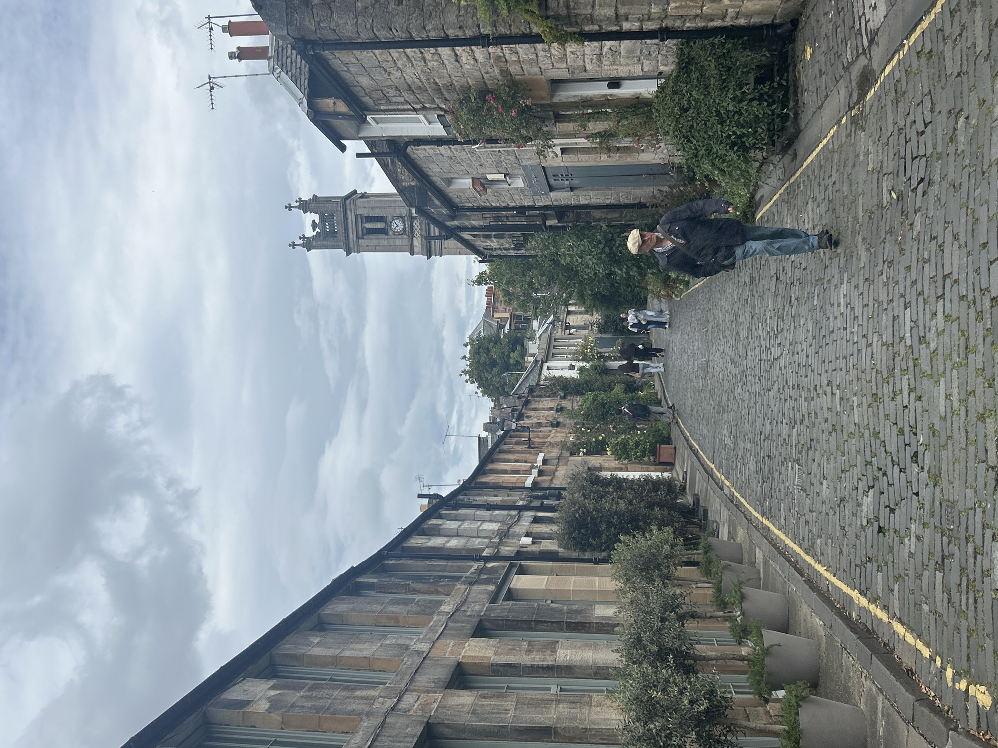 A man walks down a cobblestone street lined with stone buildings in the Stockbridge area of Edinburgh, Scotland. The man, wearing a flat cap, jacket, and jeans, walks towards the camera. Behind him, other pedestrians are visible, as well as potted plants and trees. In the background, a church tower with a clock face stands against a cloudy sky. The street is wet, suggesting recent rain.