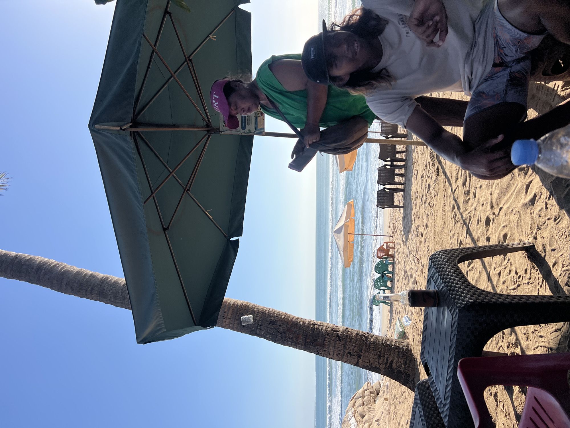 A man and a woman are sitting at a beach cafe in Kuta, Indonesia. The man is wearing a t-shirt and shorts, and the woman is wearing a green shirt and a pink hat. They are both looking at a phone. The beach is sandy, and there are several chairs and tables set up. The ocean is visible in the background, with waves crashing on the shore. A palm tree is to the left of the frame. The sky is clear and blue.