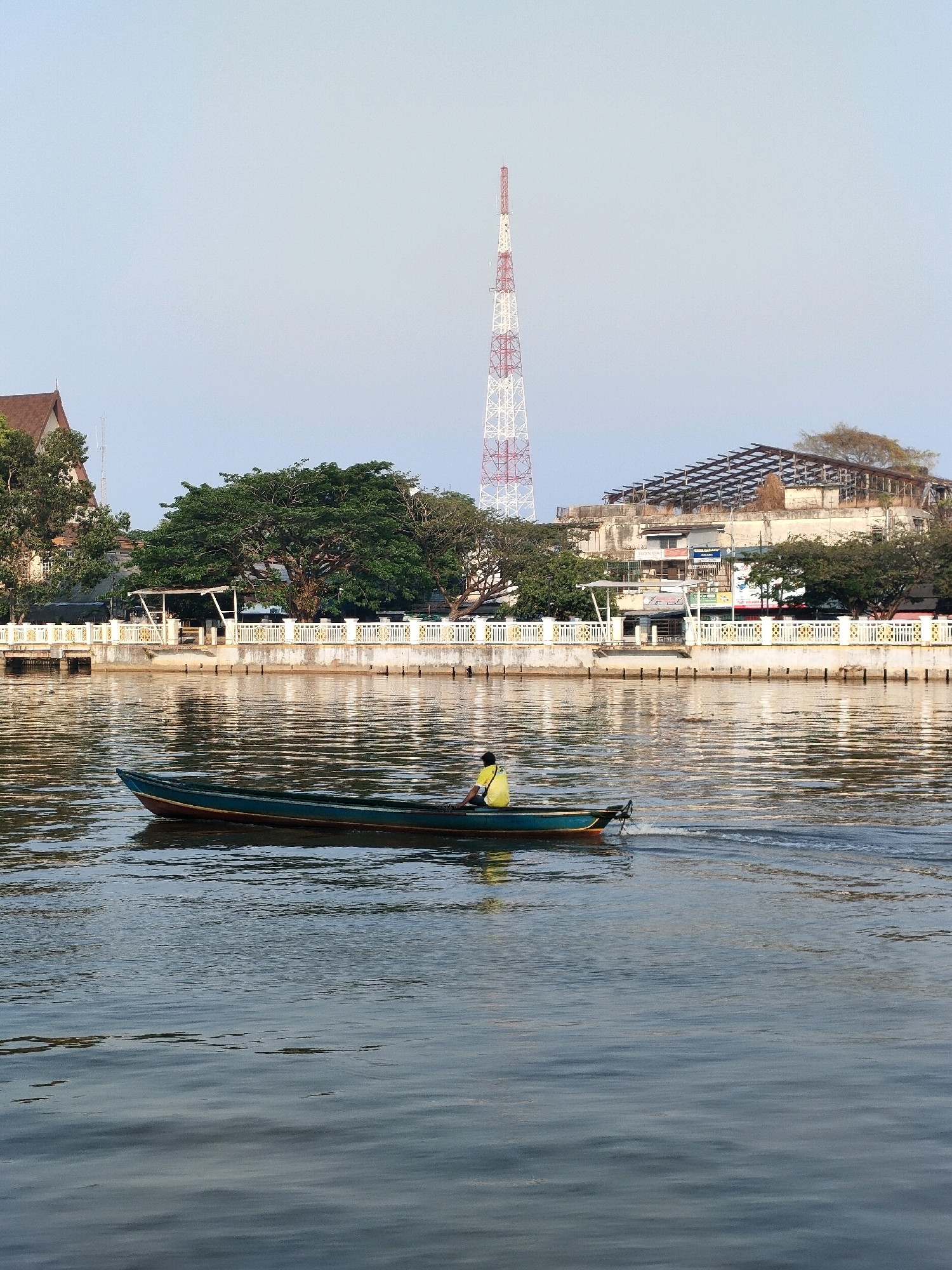 This is a medium shot, eye-level, static, outdoor, daylight, color photograph taken in Banjarmasin, Indonesia. The photograph captures a long, narrow, dark teal and red boat with a single occupant wearing a bright yellow shirt, navigating a calm body of water. The boat is positioned in the lower left quadrant of the image and is moving from left to right, leaving a visible wake. In the background, a white and red lattice tower rises prominently against a clear, pale blue sky. Along the waterfront, there is a decorative white and gold railing. Behind the railing are various buildings, including one with a dark, sloped roof and another with an exposed, unfinished roof structure. Lush green trees are scattered throughout the background, with a particularly large tree on the left side. The water surface reflects the sky and the structures along the bank, creating a shimmering effect. The overall scene depicts a tranquil moment on the river with elements of urban infrastructure present.