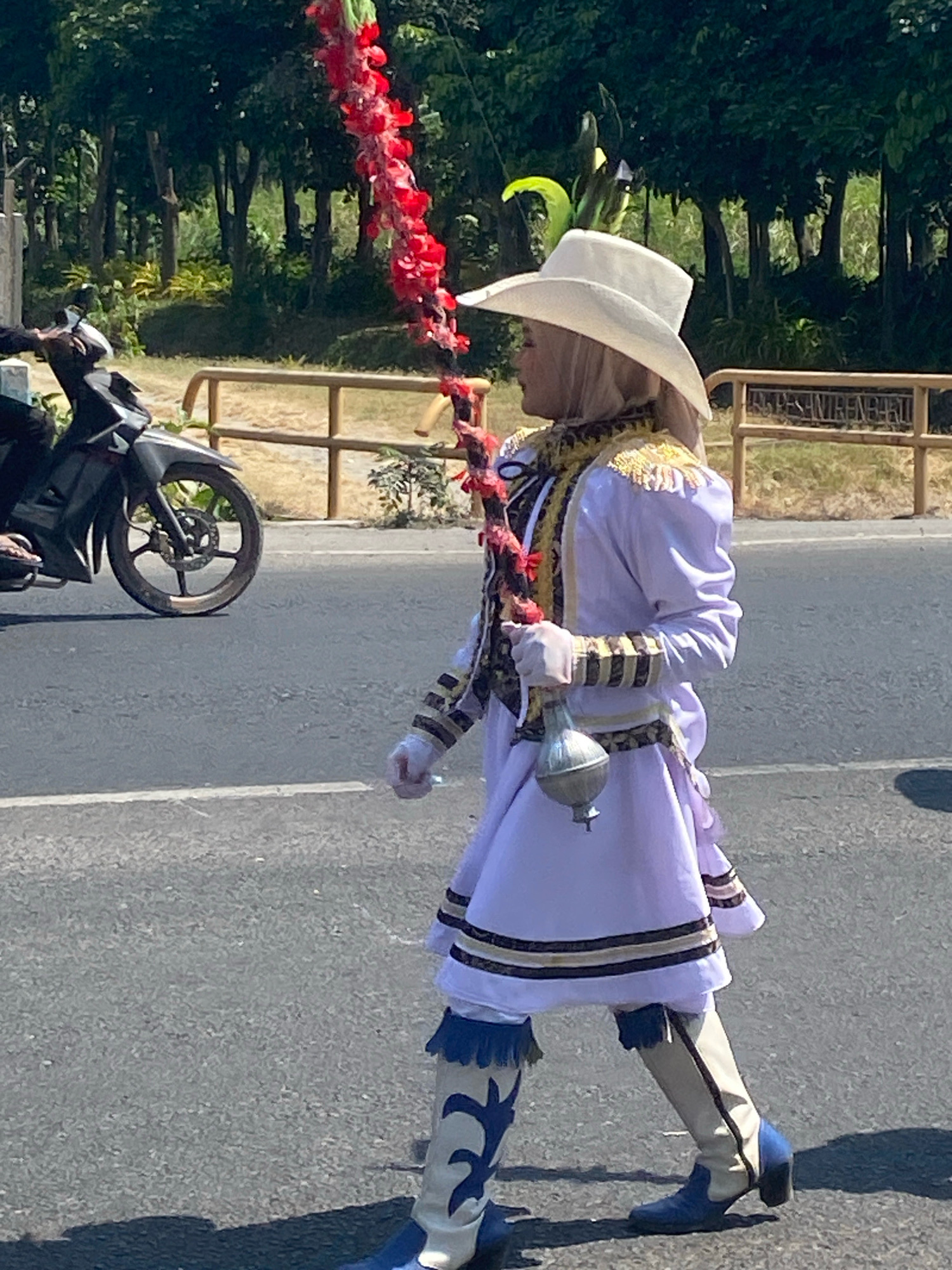 A person in a white cowboy hat and a white and gold marching band uniform with a purple skirt walks on a paved road in Ngasem, Indonesia. The person holds a long, red, flower-like object and wears white gloves and knee-high white and blue boots. A dark-colored motorcycle is parked to the left of the frame. A wooden fence with yellow posts runs along the side of the road, behind which is a dense line of trees and green foliage. The ground is asphalt with a white line marking the edge of a lane. The lighting suggests it is daytime with bright sunlight.