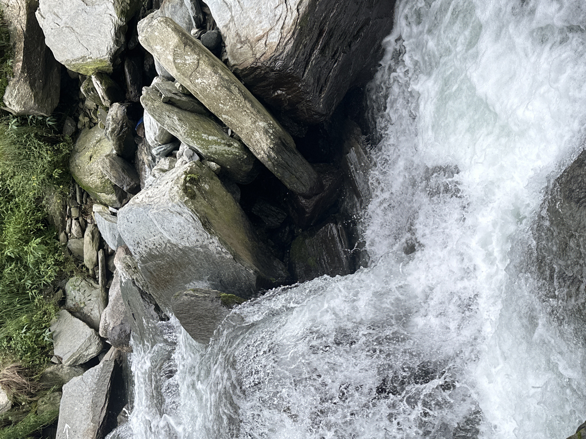 This is a photograph of a rushing stream with boulders and lush green vegetation in the background. The water is turbulent, with white foam and splashes indicating its rapid flow. Large, grey rocks of various sizes are scattered throughout the scene, some partially submerged in the water. The vegetation appears dense and grows along the banks and on higher ground. The scene is captured at an eye-level perspective, focusing on the natural elements of the environment. The location is identified as Bhagsu, India.