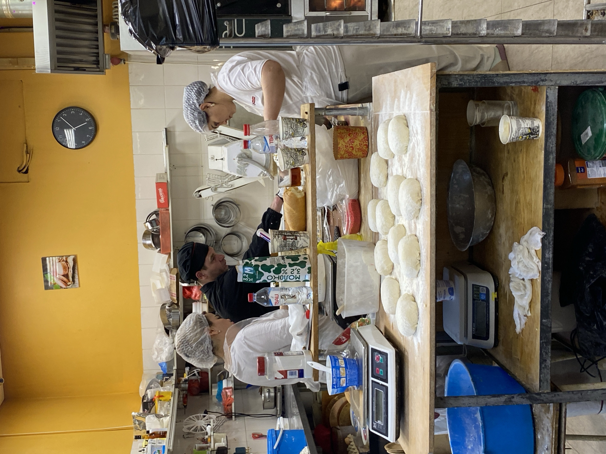The image shows a bakery in what is presumably Moscow, Russia. Two bakers wearing white uniforms and hairnets are working at a wooden counter, preparing dough. One baker is wearing a black cap. One is weighing ingredients on a scale, while the other appears to be shaping the dough into rolls. A shelf behind the counter holds various baking supplies and equipment, including bowls, bottles, cups, and canisters. The walls are painted yellow, and a clock hangs on one wall.