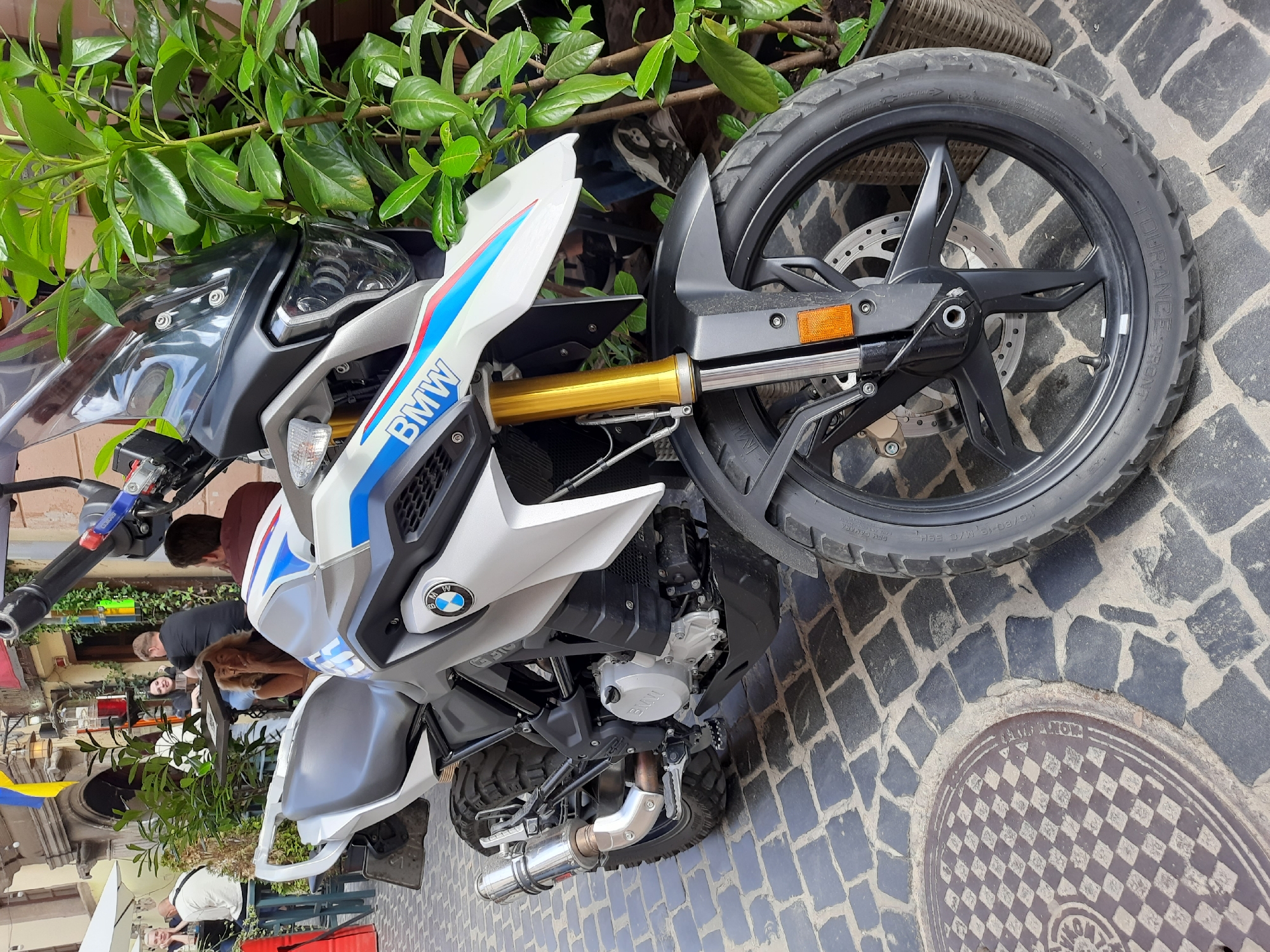 A silver and white BMW motorcycle with red, white, and blue racing stripes and "BMW" branding is parked on a cobblestone street in Lviv, Ukraine. The motorcycle has knobby tires, suggesting it's an adventure or dual-sport model. A windshield is mounted on the front, and golden-colored front suspension components are visible. The motorcycle is positioned next to a potted plant with lush green leaves. In the background, several people are seated at tables outside a cafe or restaurant. Some buildings with awnings are also visible, and a Ukrainian flag is hanging from one of them. The scene appears to be a daytime shot on a pedestrian-friendly street.