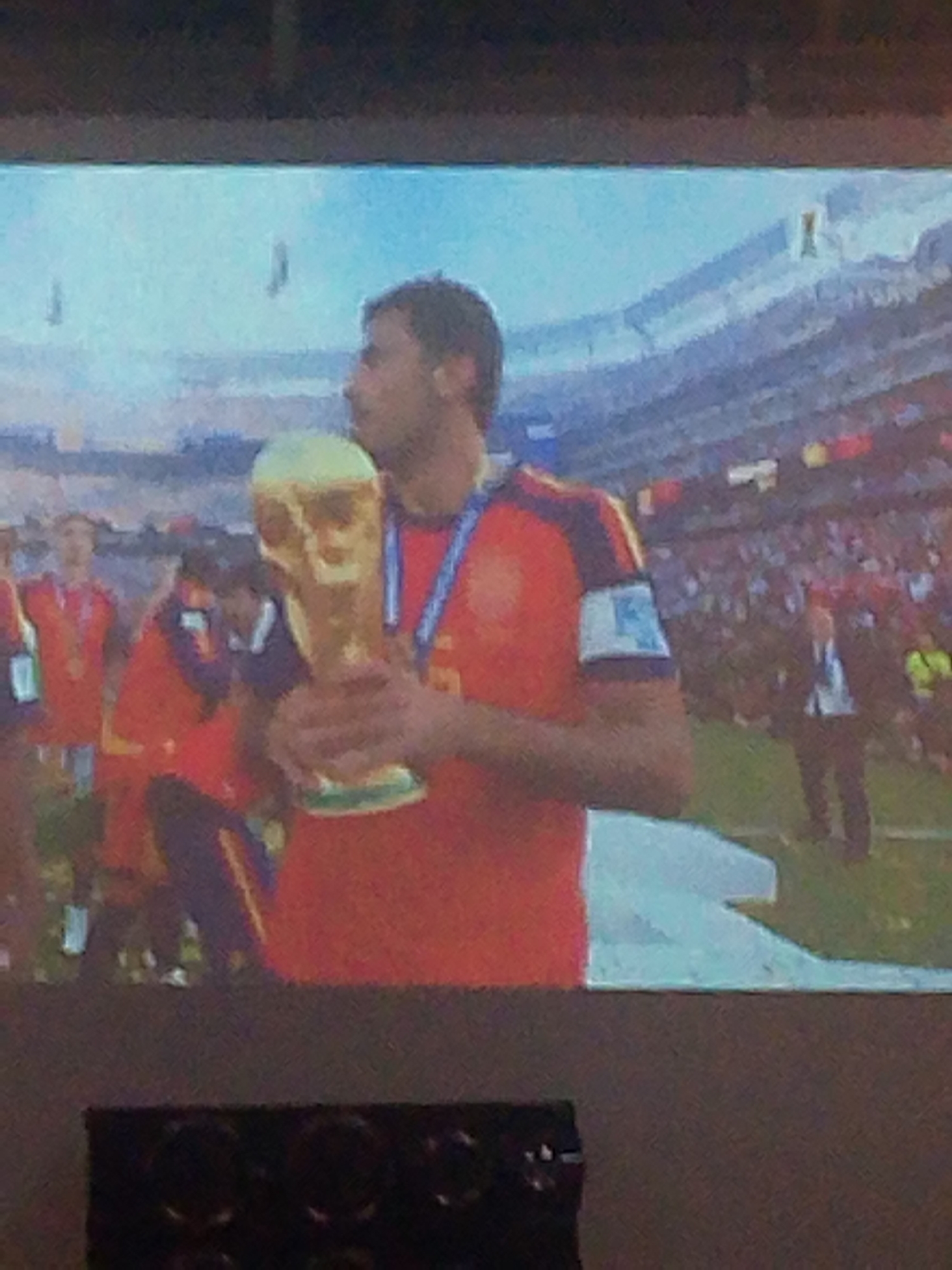 The image displays a projection of a soccer player holding a golden trophy, with a blurred stadium and spectators in the background. The player is wearing a red jersey with blue and yellow trim. A man in a suit is visible in the lower right quadrant of the projected image, possibly an official. The lower portion of the image shows a dark, out-of-focus object that could be a piece of furniture or equipment. The lighting conditions suggest an indoor viewing of a projected image. There is no discernible information within the image to indicate the location as Meunasahmee, Indonesia.