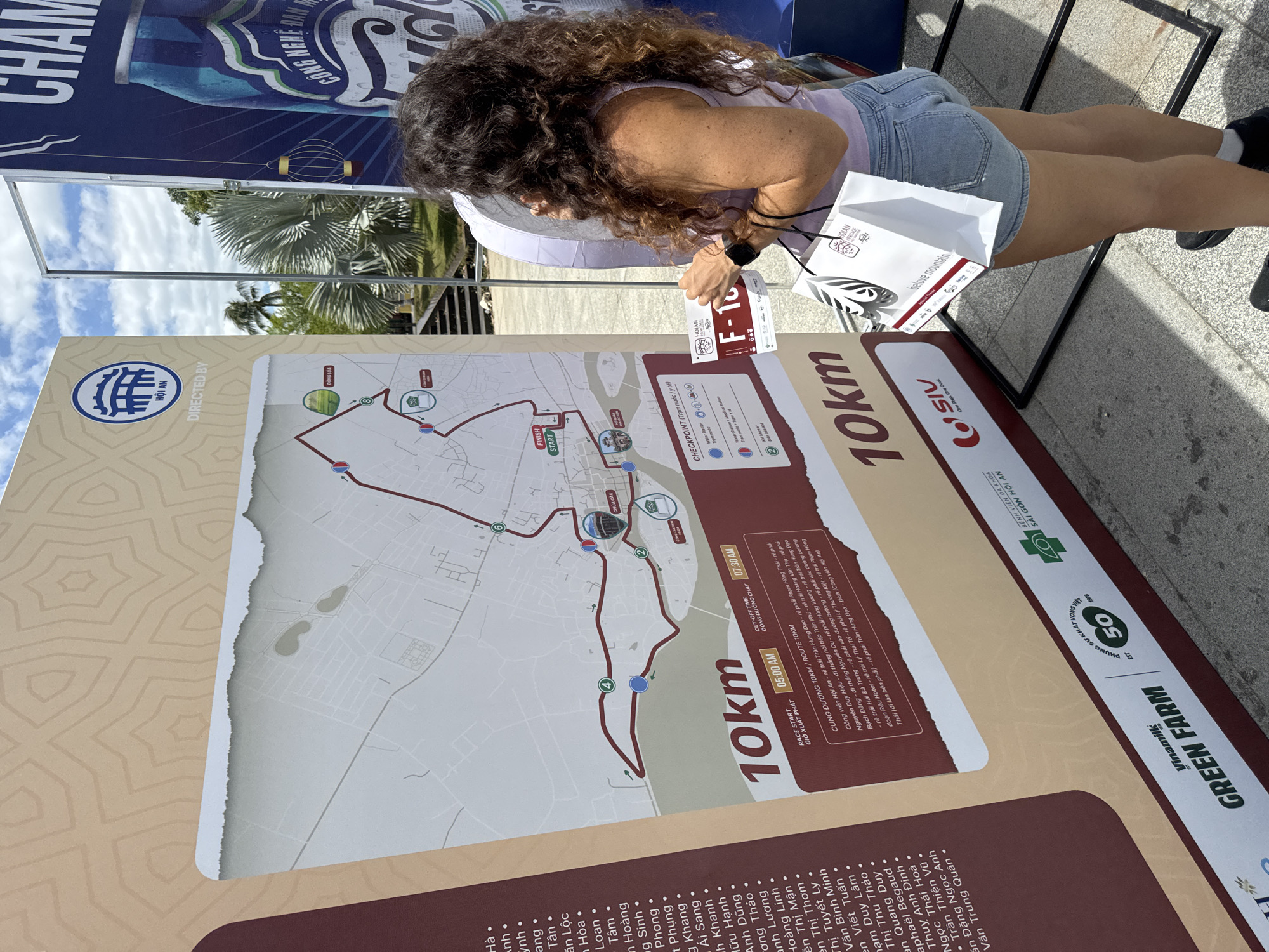 A woman with long, curly brown hair stands in front of a large, tan and brown informational display. The display features a map of a running route, with a red line indicating the course. Text labels on the display include "10km", "RACE START 05:00 AM", and "CUT-OFF TIME 07:30 AM". There are also Vietnamese inscriptions and logos. The woman is wearing a purple top and denim shorts, and she is holding a white paper bag with a Hoi An Heritage logo and a bib with "F-16" printed on it. To the right of the display is a tall, dark blue banner with "CHAMPION" and "CÔNG NGHỆ ĐAN MẠCH" printed on it, next to an image of a bottle. In the background, palm trees and a bright blue sky with white clouds are visible. The ground appears to be paved with stone. The scene is outdoors, suggesting an event or festival in Hoi An, Vietnam.