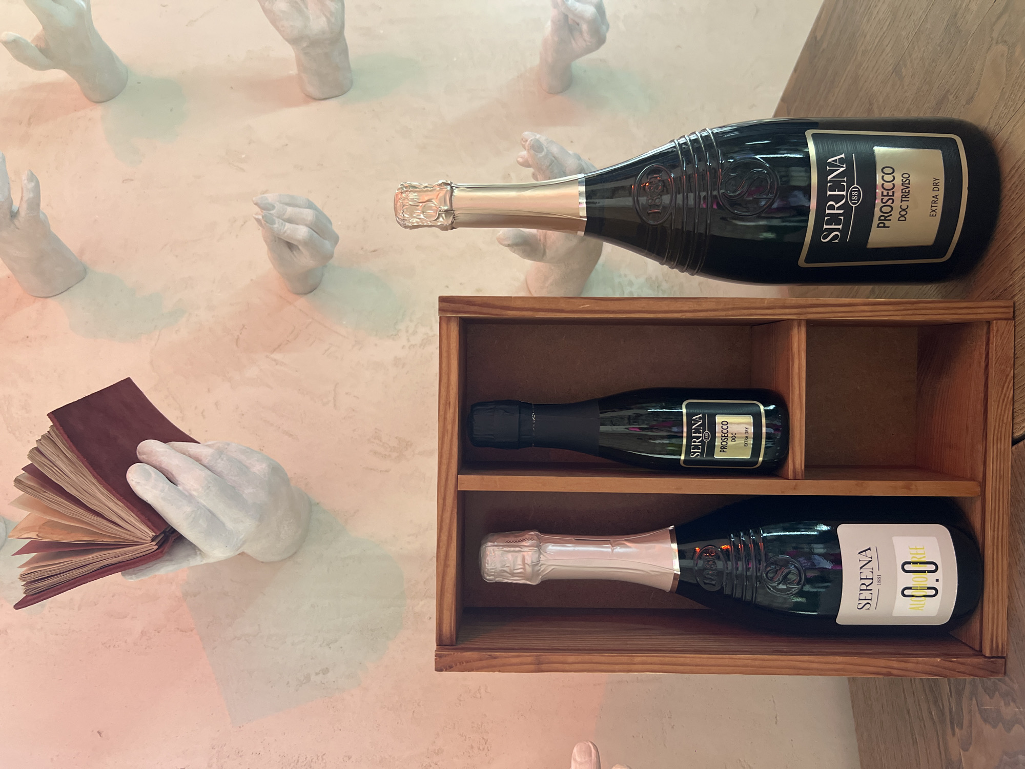 This image depicts a still life setup with several bottles of Serena brand Prosecco and an artistic wall decoration featuring plaster casts of hands emerging from a textured wall. In the foreground, a wooden shelf unit holds two bottles of Serena Prosecco. The bottle on the left is a larger size and is labeled "SERENA 1881 ALCOHOL FREE 0.0". It has a silver foil cap. The bottle on the right is a smaller, mini-sized bottle and appears to be a standard Prosecco DOC, also with a Serena label and a black foil cap. To the right of the shelf unit stands a larger bottle of Serena Prosecco DOC Treviso, labeled "EXTRA DRY", with a gold foil cap. The background wall is a pale, textured surface with several white plaster casts of hands. One hand, in the upper left, holds open a small, dark red book. Other hands are positioned as if reaching or gesturing. The lighting suggests an indoor setting, possibly a restaurant or bar. The location is identified as Bender, Moldova, though there are no specific landmarks in the image to confirm this.
