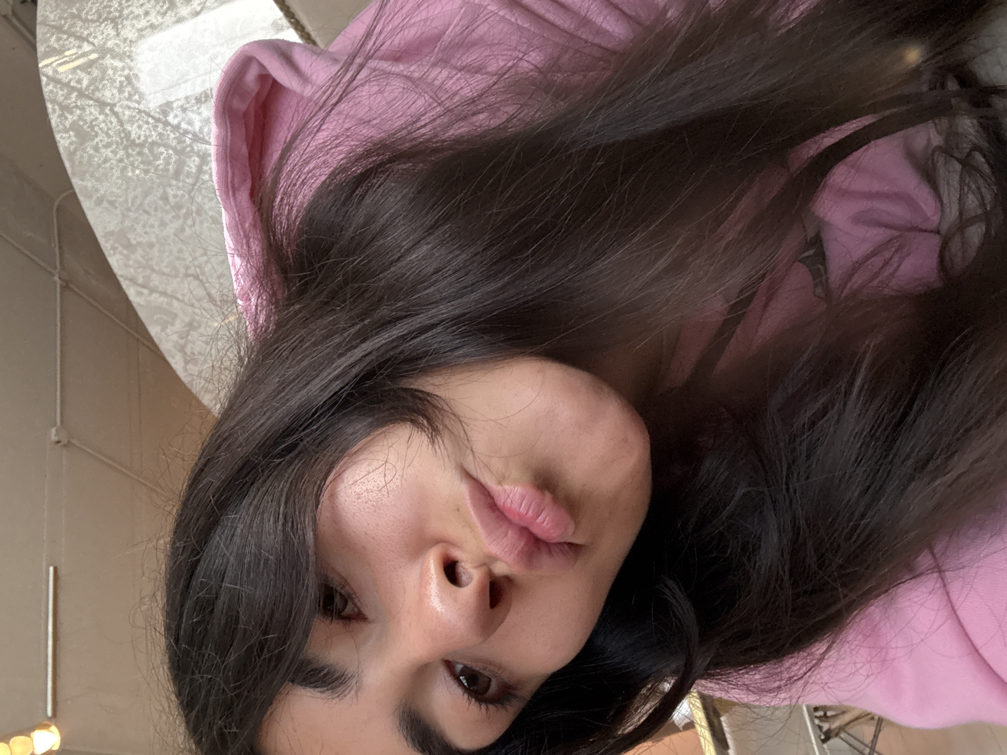 The image is a close-up selfie of a person, likely female, with long, dark, slightly wavy hair framing their face. The subject is wearing a pink hooded sweatshirt, parts of which are visible around their head and behind their hair. Their head is tilted, and they are looking towards the camera with a playful expression, forming a "kissy face" or pout with their lips. Their features include dark eyes, well-defined eyebrows, and a small nose.
The background indicates an indoor setting, characteristic of a commercial or public space such as a cafe or restaurant in Puebla, Mexico. To the upper left, there is a bright, out-of-focus area, possibly a window or a reflective surface, revealing intricate, tree-like patterns or reflections. Further left and at the bottom, a light, neutral-colored wall or pillar is visible, along with out-of-focus warm lights. The lighting suggests it is daytime. The overall mood is casual and expressive. There is no visible text in the image.