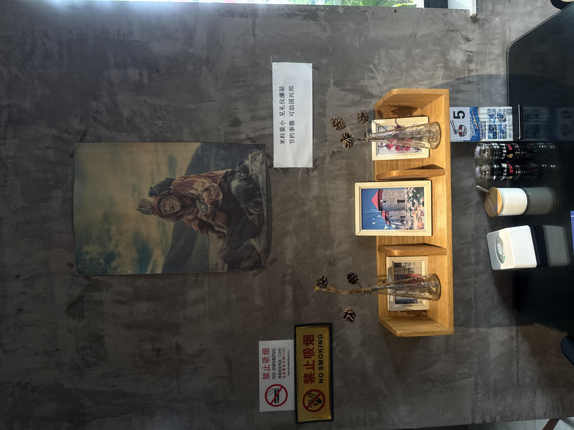 The image depicts an interior scene featuring a textured concrete wall adorned with several items. Prominently displayed is a framed poster of an elderly person playing a guitar in a mountainous landscape. To the left of the poster, two "NO SMOKING" signs are visible, one red and white, and another with a yellow border. Below these signs, a wooden shelf is mounted on the wall, holding three framed pictures of buildings, two glass vases with pine cones, and a bottle of flowers. On the table below the shelf, there are two jars of condiments, a tissue dispenser, and a menu with a prominent number "5". The overall ambiance suggests a cafe or a restaurant setting. The specific location in Qinhuangdao, China, cannot be determined from the visual information provided in the image.
