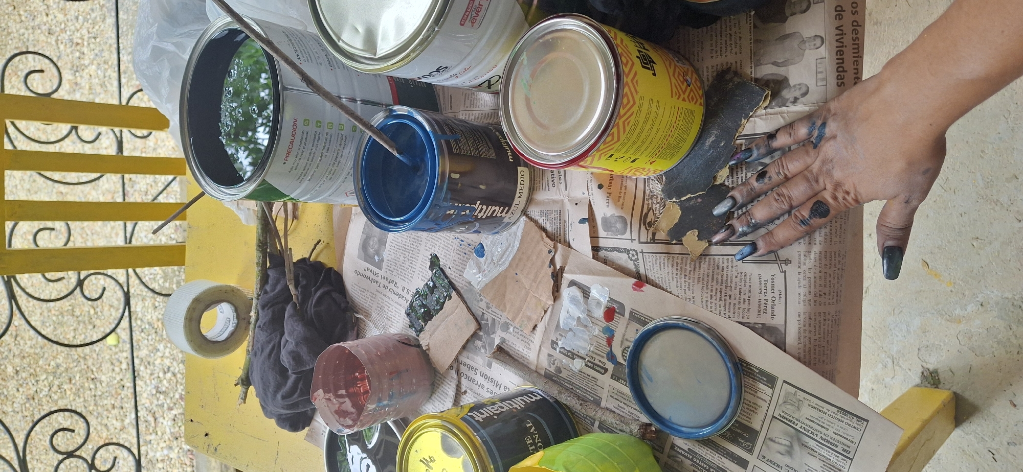 This is a top-down, close-up shot of a cluttered yellow table, likely outdoors given the blurred background of a yellow metal railing and gravelly ground. The scene is a DIY painting setup. In the center, a hand with long, dark, painted nails, smudged with dark paint, rests on a spread of newspapers. The hand is positioned over a piece of dark sandpaper and some torn cardboard.
Scattered around the table are several cans of paint. A large green can with a white label that says "PRECAUCION" is partially visible at the top right. Next to it, a white can, also with a label, is present. A blue can, labeled "MULTIPURPOSE AZUL MEDIO," is in the middle, with a paint stirrer inside. To the left, a yellow can, labeled "MULTIPURPOSE," is visible. Another can or container, possibly plastic and painted green, is at the bottom left. A smaller, red-painted plastic container is positioned between the green and blue paint cans.
Also on the table are a roll of white tape, some dried sticks or twigs, and a piece of dark cloth. The newspapers serve as a protective layer, showing various articles and advertisements. Splatters of paint, predominantly blue and some red and white, are visible on the newspapers, cardboard, and even on the hand. The overall impression is one of an ongoing, somewhat messy, painting project. The location in Vallecito, Venezuela is not indicated by any visual cues within the image itself.