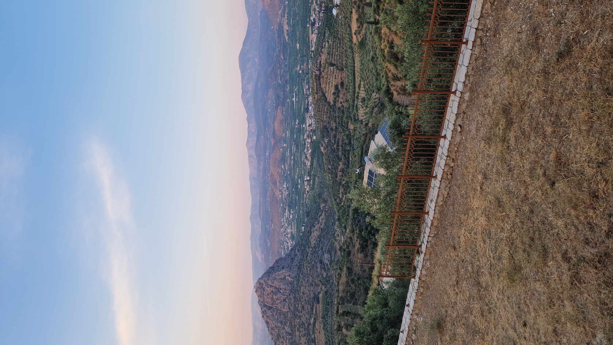This is a vertical, eye-level, outdoor, daytime photograph taken in the Municipal Unit of Asini, Greece. The scene depicts a panoramic view of a valley dotted with buildings and verdant hills. In the foreground, a dry, grassy slope is bordered by a rustic, rusty metal fence. Beyond the fence, lush green vegetation, likely olive trees, covers the rolling hills. A large, rocky mountain with a structure on its summit dominates the left side of the frame. In the mid-ground, a cluster of buildings is nestled within the valley, and a few buildings with solar panels are visible on a hillside. The background features a range of hazy, purple mountains under a clear, pale blue sky with a wispy white cloud. The overall impression is one of a peaceful rural landscape at dusk.