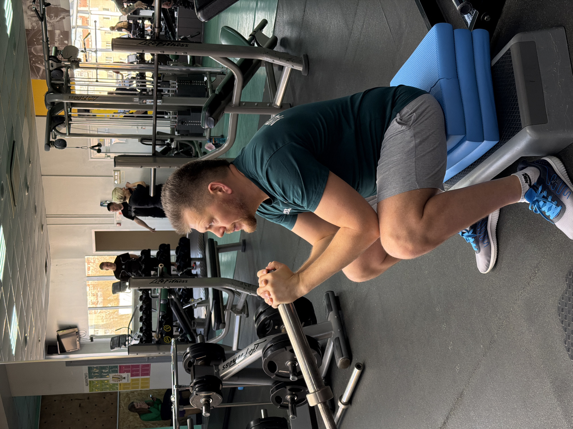 The image shows a man in a gym. He is wearing a dark green t-shirt, gray shorts, and blue and white athletic shoes. He is using a machine, positioned with his knees on a blue padded support and leaning forward, gripping a silver bar with both hands. The machine has black weight plates loaded on it.
In the background, other gym equipment and people are visible. Through a window, a building is seen. Based on the building visible through the window and the presence of Life Fitness equipment, the location is likely a gym in Izhevsk, Russia.