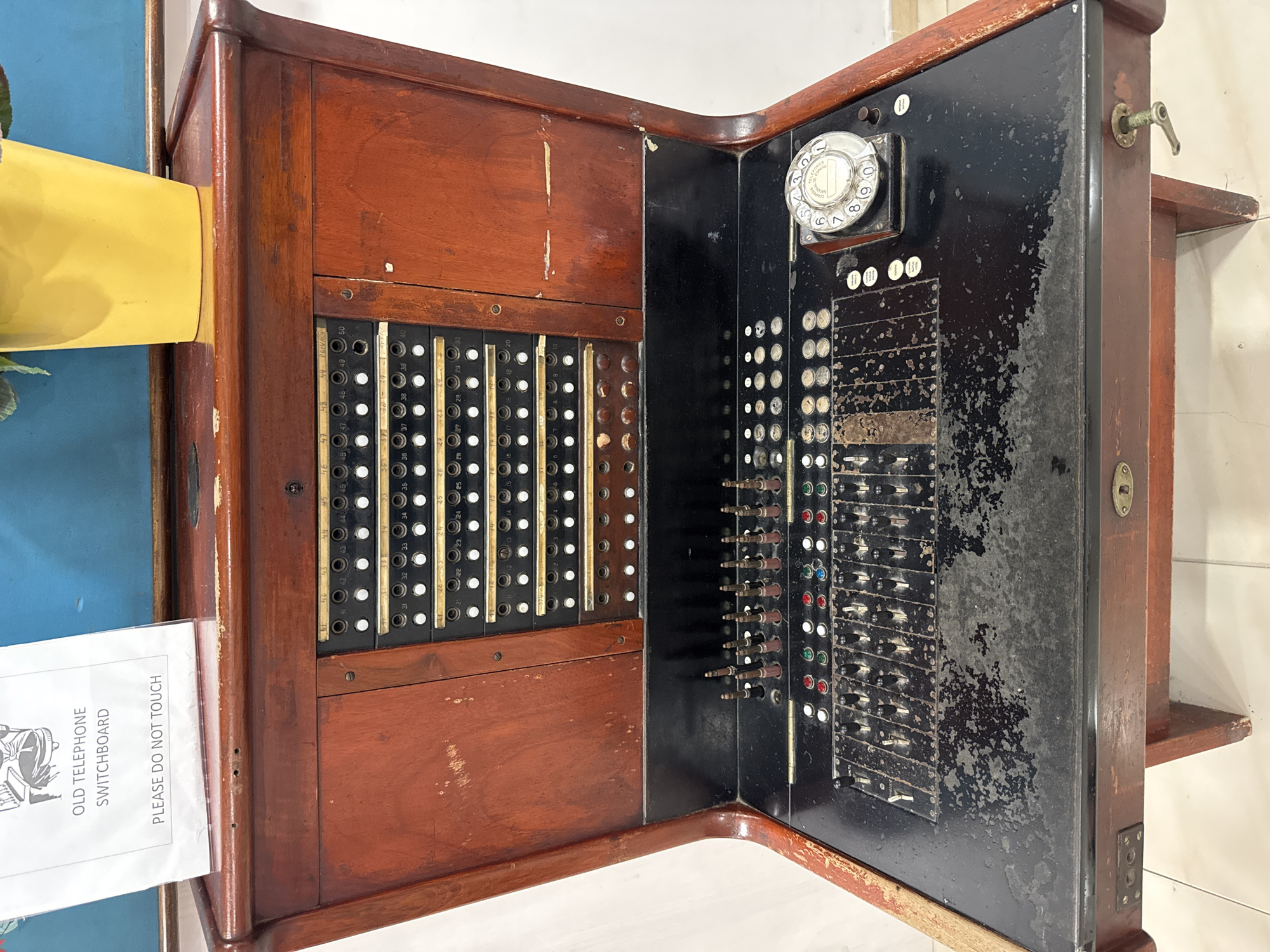 An antique wooden telephone switchboard is displayed in a room in Can Pastilla, Spain. The switchboard is a large, upright wooden console with a black surface featuring numerous switches, buttons, and jacks. A circular rotary dial is visible on the right side of the black surface. Above the black surface, a panel with many round buttons labeled with numbers is visible. A sign that reads "OLD TELEPHONE SWITCHBOARD PLEASE DO NOT TOUCH" is placed on the top left of the wooden console. A yellow potted plant sits on the right side of the top of the console. The background of the image is a blue wall. The switchboard is the sole subject in the image.