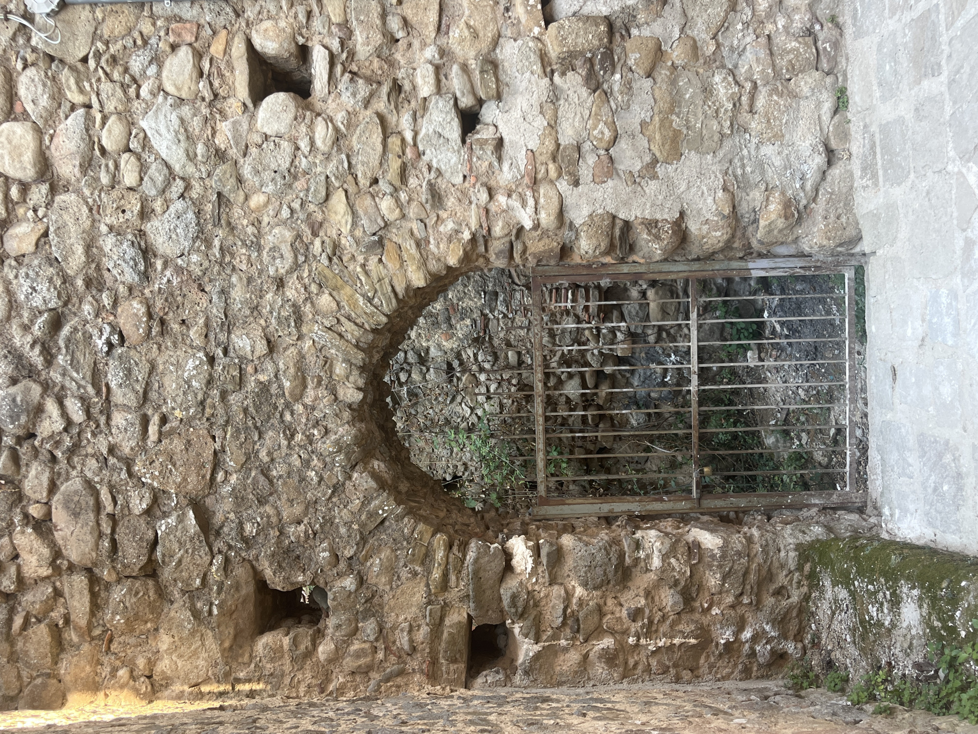 This image depicts a rustic stone archway with a weathered metal gate in Vallo della Lucania, Italy. The stonework of the archway is rough and uneven, composed of various sizes and colors of natural stones, interspersed with patches of older mortar. Two small, square openings are visible in the stonework to the left and right of the arch, suggesting historical defensive features or ventilation shafts. The metal gate is simple, with vertical bars and a small padlock securing it. Beyond the gate, dense greenery and more stonework are visible, hinting at a hidden courtyard or passage. The ground in front of the arch is paved with light-colored, irregular flagstones, with a moss-covered stone ledge to the left. The overall scene conveys a sense of age and neglect, characteristic of historical architecture in older European towns.