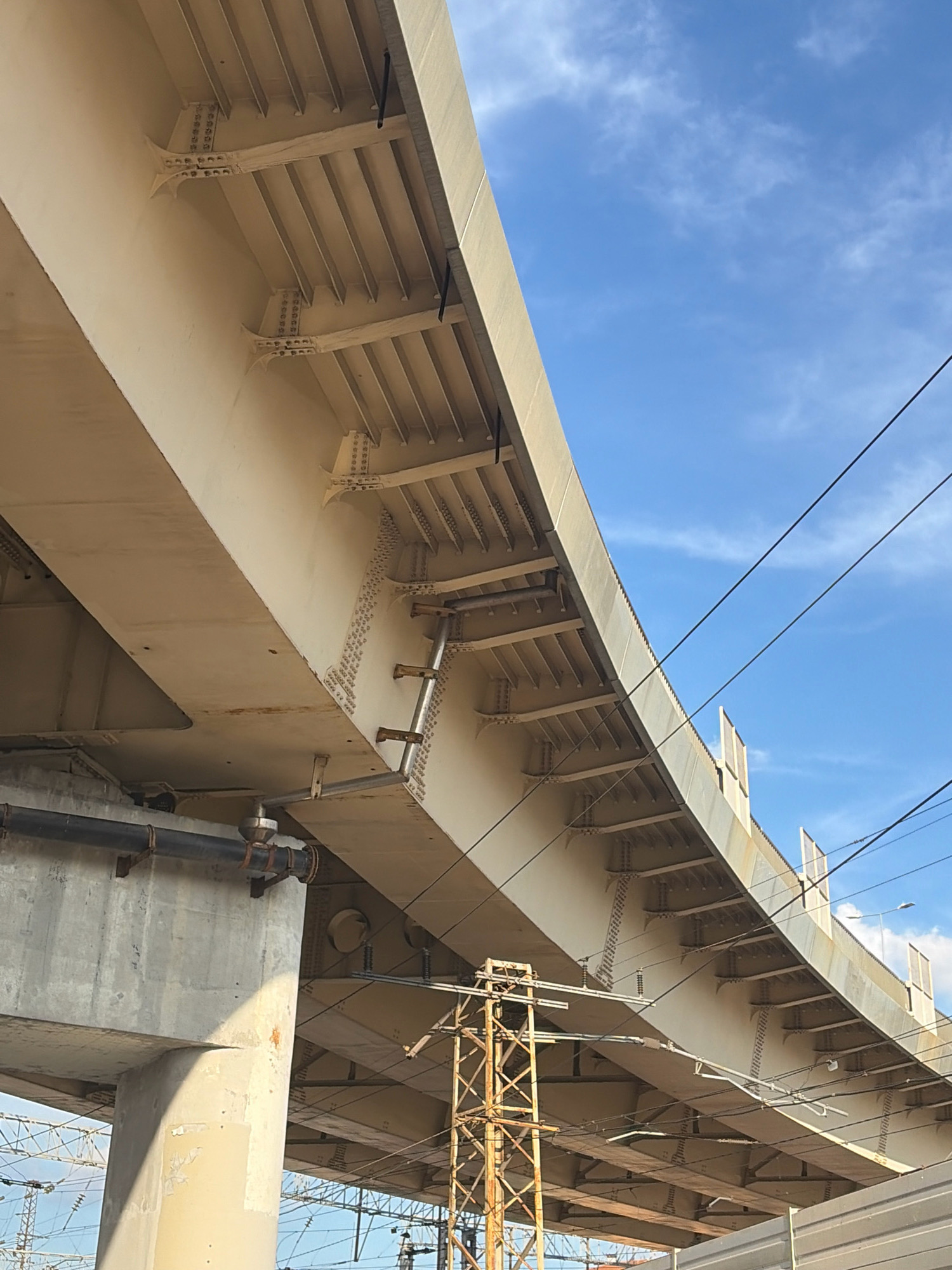 The image depicts the underside of an elevated road or bridge structure, viewed from below against a bright blue sky with scattered white clouds. The structure is a light beige color and appears to be made of concrete and steel beams. Several vertical steel beams are visible supporting the horizontal structure, with numerous rivets connecting various components.
Beneath the main structure, a large concrete pillar supports one side. A thick black pipe, likely for utilities, is attached to the underside of the concrete pillar with metal brackets. In the background, a brown, lattice-like metal tower, potentially a utility pole or part of the bridge's infrastructure, rises up. Further in the distance, power lines and insulators are visible, suggesting an urban or semi-urban environment, possibly a transportation hub. The overall scene conveys a sense of large-scale infrastructure. The context provided indicates this location is in Nizhny Novgorod, Russia, suggesting this is an overpass or bridge within the city.