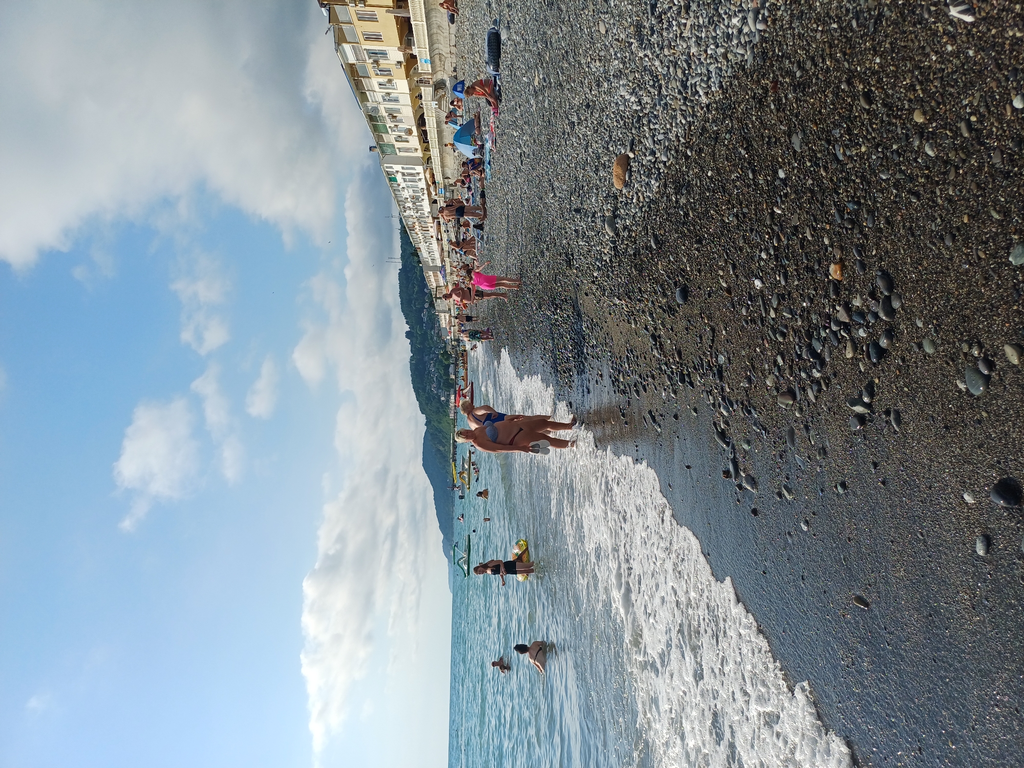 This is a photograph of a crowded beach in Sochi, Russia. The image is oriented vertically, with the sea on the left and the beach and buildings on the right. The sea is a clear blue with gentle waves lapping at the shore. Several people are in the water, some swimming and others wading.
The beach itself is made of dark pebbles. People are scattered across the beach, sunbathing, sitting, and walking. There is a row of buildings behind the beach, which appear to be hotels or apartment blocks, with balconies overlooking the sea. A green, tree-covered mountain can be seen in the background, rising behind the buildings.
The sky is a bright blue with scattered white clouds. The sun is shining, suggesting it is a warm, pleasant day, likely during the summer months. The presence of many people in swimwear and the overall atmosphere of leisure indicate a holiday or recreational setting. There is no visible text in the image.
