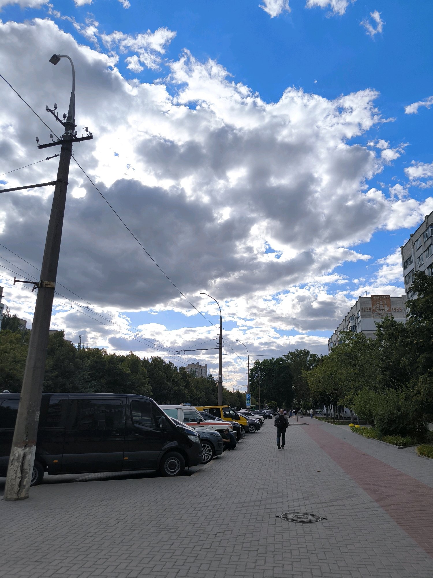 This is a medium shot, taken from a sidewalk, of a street scene with parked vehicles and a pedestrian walking away from the camera. The sky is a vibrant blue with large, fluffy white and gray clouds. In the foreground, a dark-colored van is parked on the left. To the right of the van, other vehicles, including what appears to be an ambulance and a yellow van, are parked in a row. The sidewalk is paved with gray interlocking bricks, with a pink and red striped section running alongside it on the right. A single pedestrian, wearing dark clothing and a backpack, is walking away from the viewer down the sidewalk. Trees line the right side of the street, and apartment buildings are visible in the background. A large utility pole with electrical wires is prominent on the left side of the image. This scene is located on General Petrov Street in Mykolaiv, Ukraine, as indicated by the presence of the "MYKOLAIVENERGO" building in the background, which is identifiable by the text and emblem on its facade. The street has a parking area adjacent to the sidewalk.