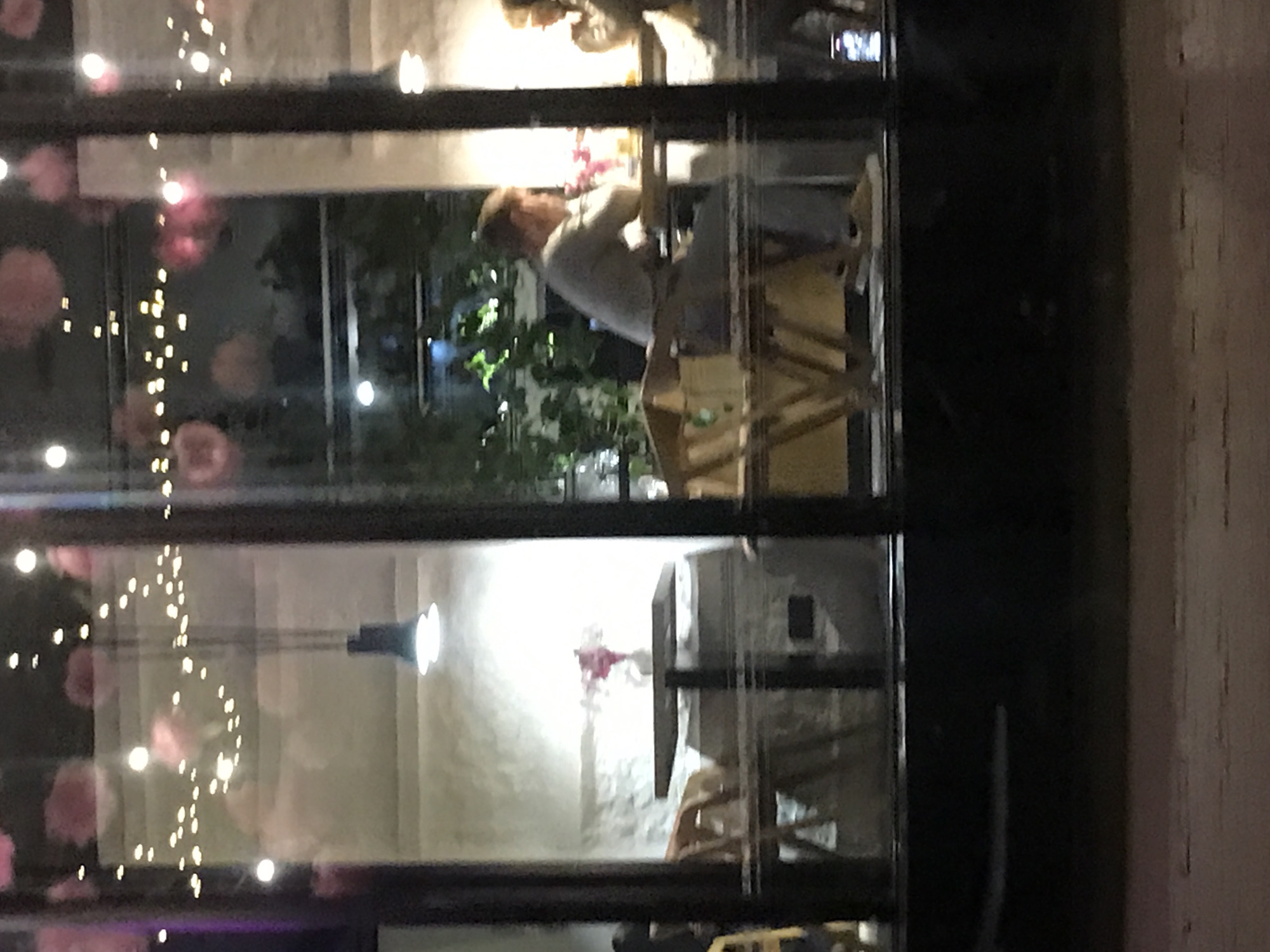 The image shows a view through a window, possibly of a café or restaurant at night. The location is unknown. String lights with pink floral accents are visible on the left side of the window. Inside, a person is sitting at a wooden table with a chair, appearing to be working or reading. A plant is located near the person, and there is a table with a floral arrangement or centerpiece in the background. A hanging lamp is visible on the lower left. The overall atmosphere seems relaxed and dimly lit.