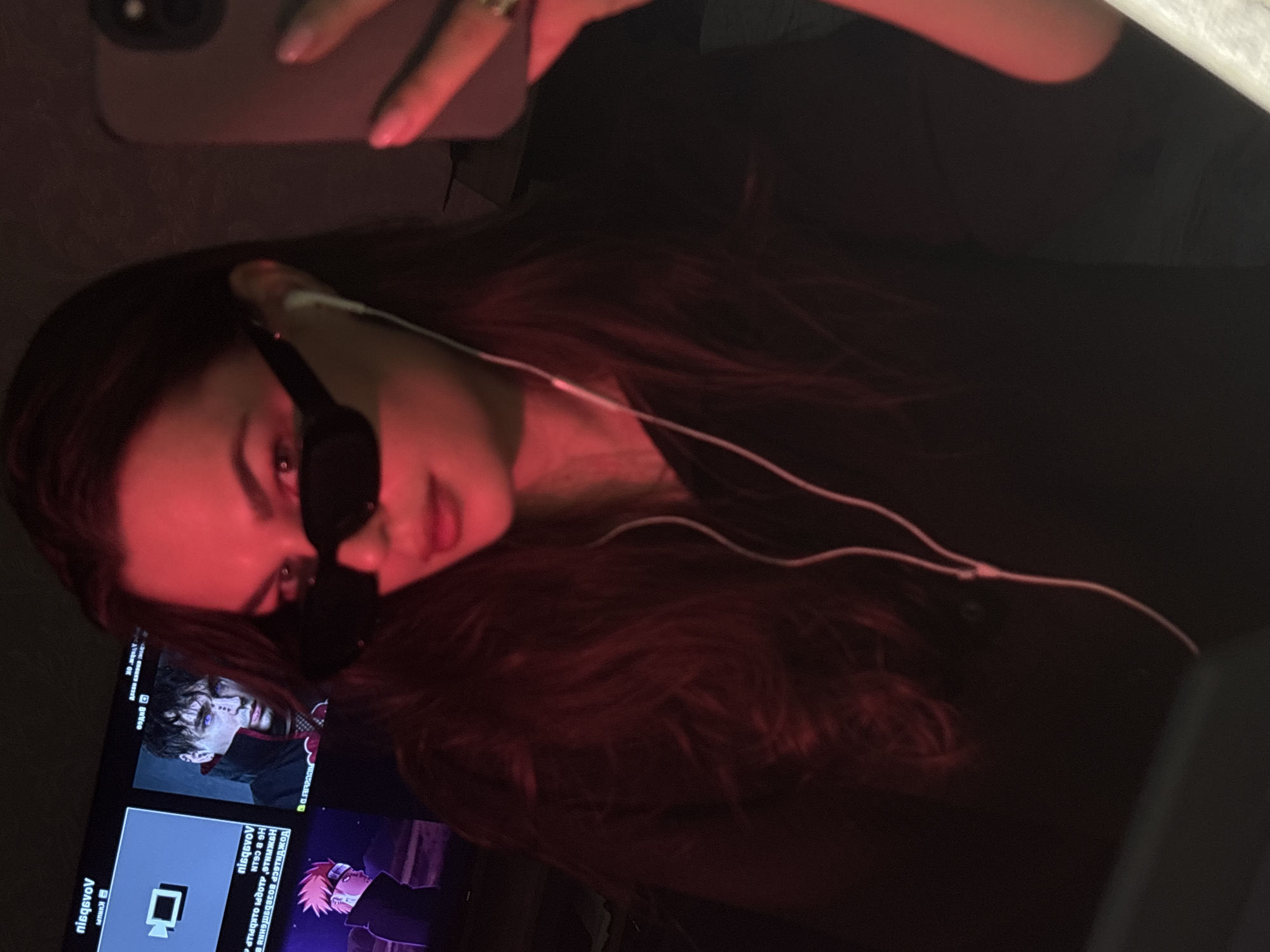 A woman with long brown hair is taking a selfie with her phone. She is wearing sunglasses and has white earbuds in her ears. The background is dark, but a television screen is visible showing images from the anime series "Naruto." The scene is bathed in a red light. There is no indication of the city of Chaikovsky, Russia.