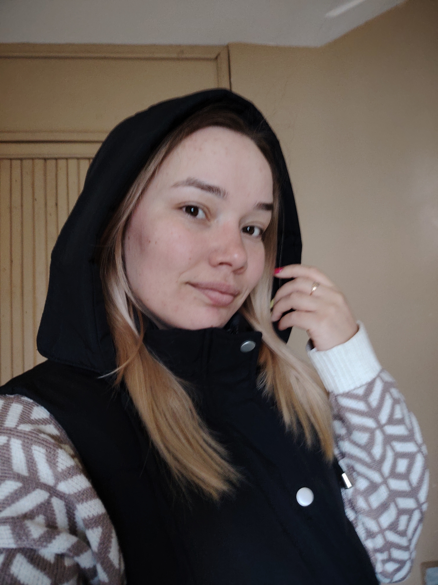 A young woman is posing for a selfie indoors. She wears a black hooded vest with a visible button and a patterned sweater. She is wearing a ring on her finger. The walls are plain and beige with a wooden section near the top. There is no specific information to confirm that this image was taken in Марковский, Russia.