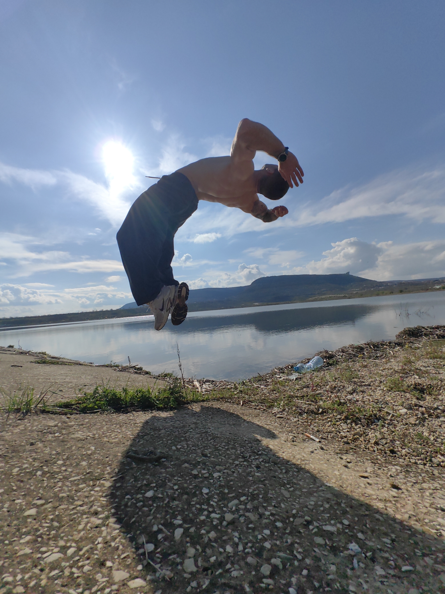 A man performs a backflip outdoors on a sunny day near a body of water. He is shirtless, wearing dark pants and white sneakers. The location appears to be a lake or reservoir, with a shoreline of dirt, grass, and some litter. A road or paved area is visible in the foreground. In the background, there are hills or mountains under a partly cloudy sky.
