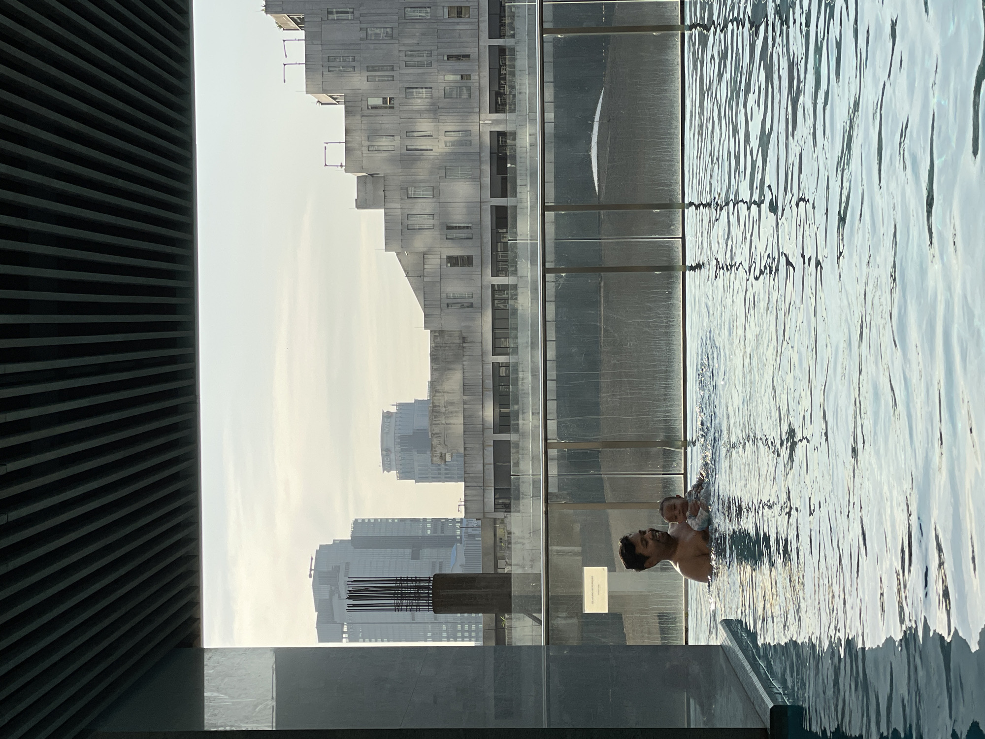 A man holds a baby in the water of an outdoor pool, with city buildings visible in the background. The location is Central Jakarta, Indonesia.
The pool is bordered by glass panels. In the background are grey concrete buildings, some partially obscured by the glass panels. One building appears unfinished with exposed rebar. A dark, slatted structure is on the left. The water is rippled, and there is a reflection of the building on the glass. The sky is hazy.