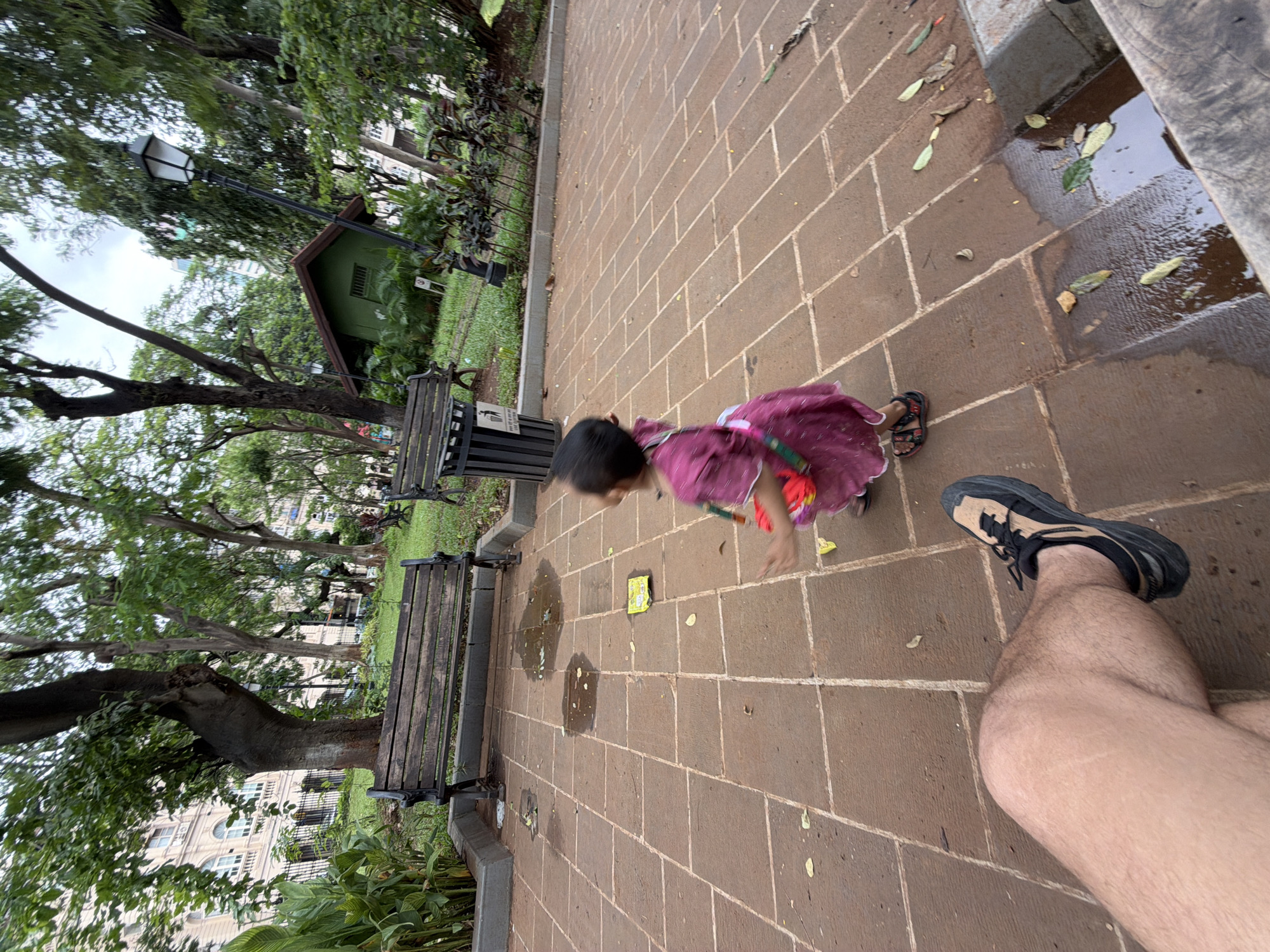 In a Mumbai park, a child in a pink dress plays on a paved ground littered with leaves. A man's legs and foot, wearing a black and tan sneaker, are in the foreground. Benches and trees are visible in the background.