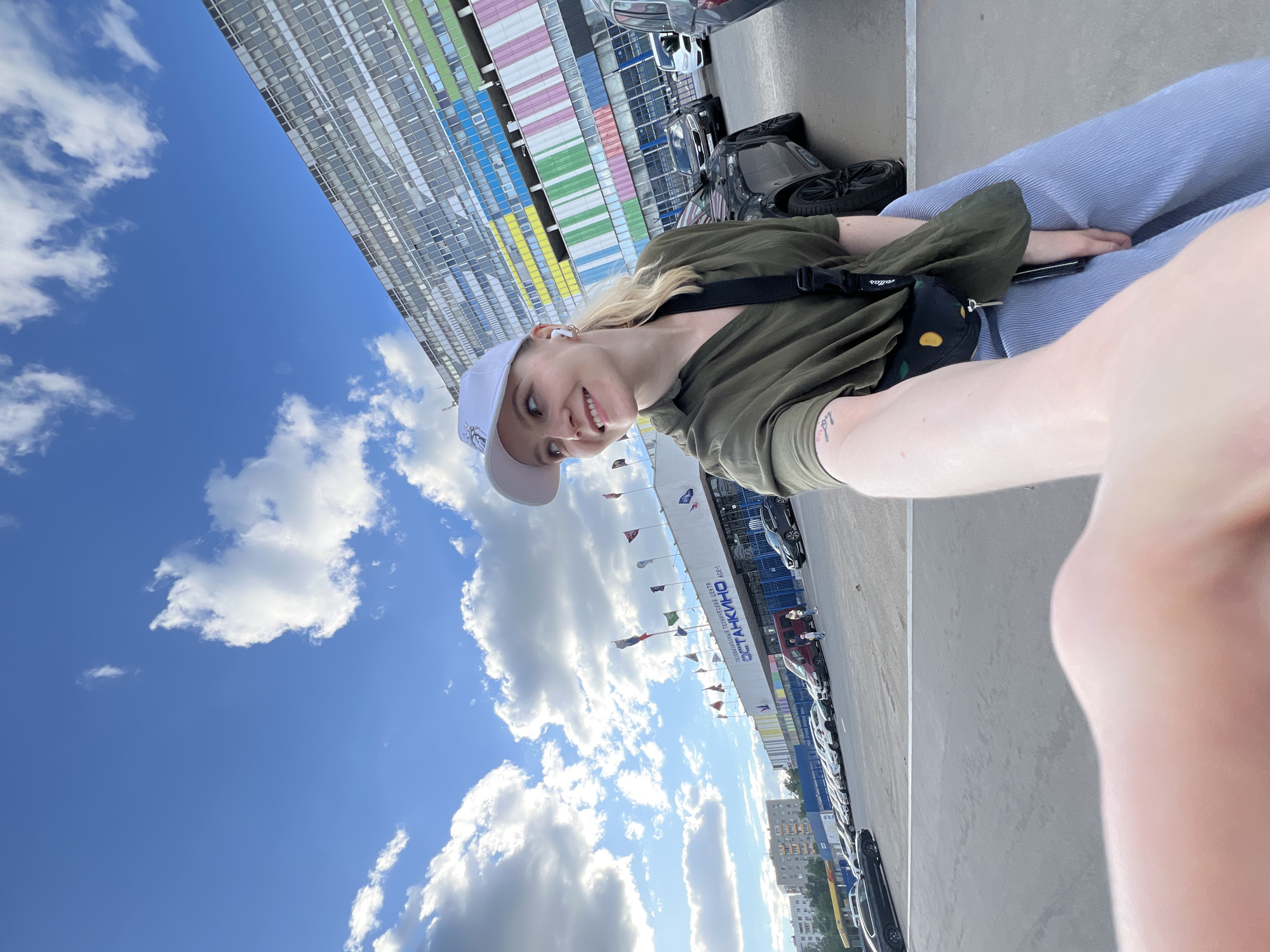 A selfie taken outdoors in Khimki, Russia, shows a young woman wearing a white baseball cap, a green shirt, and blue pants. She is seated, possibly on a ledge, with a small black bag across her shoulder. The background includes a building with multi-colored panels (pink, blue, yellow, green) and a blue sky with scattered clouds. Flags are visible in the distance. Cars are parked in rows along a paved area. A banner with Cyrillic text is seen overhead.