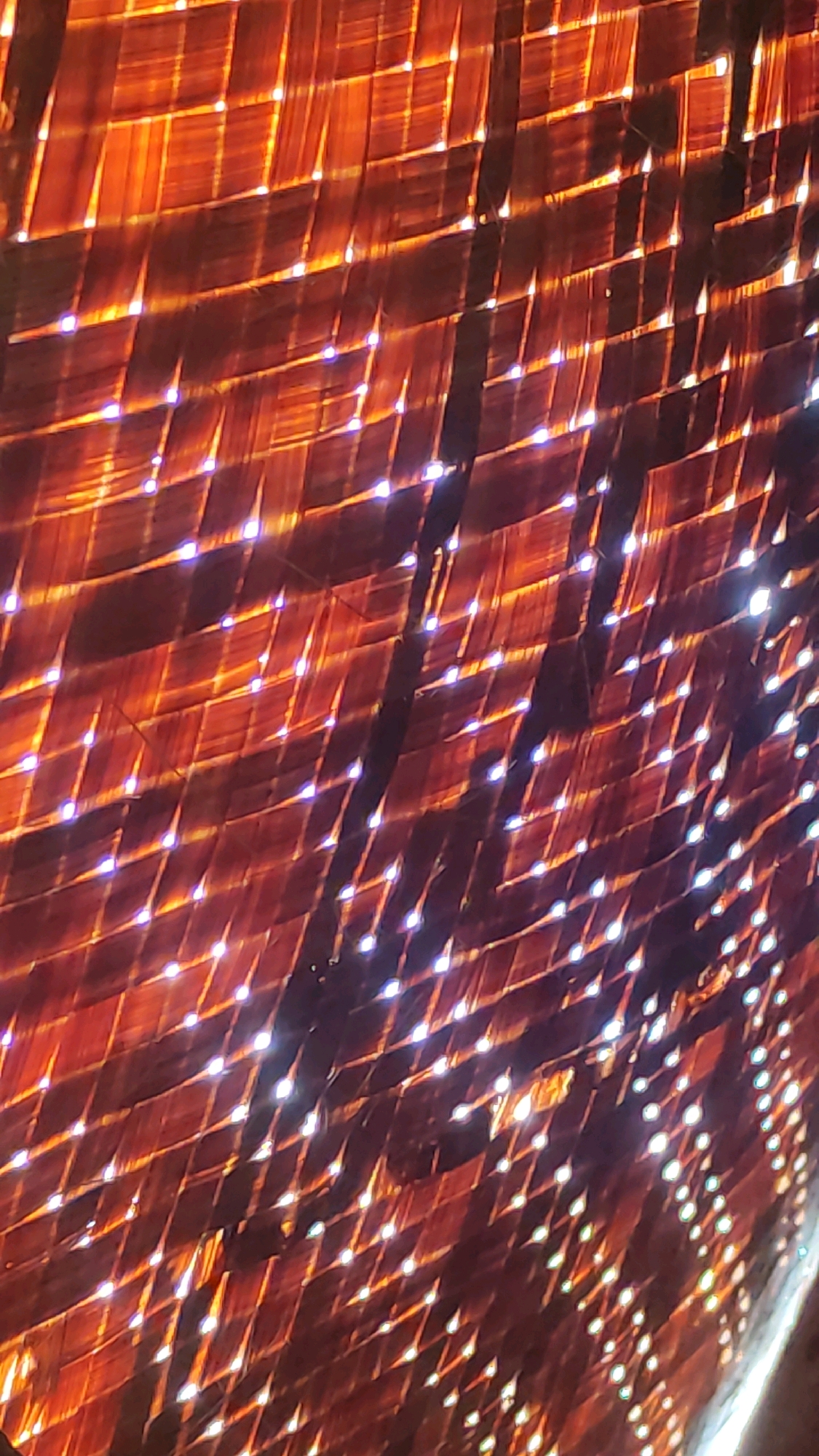 The image is an abstract close-up of a woven material, possibly bamboo or dried leaves, illuminated from behind. The texture is rough and fibrous, with individual strands clearly visible. The lighting creates a grid-like pattern of bright, white highlights interspersed with dark shadows, giving it a dappled or translucent effect. The overall color palette is dominated by warm oranges and browns, suggesting a natural material. There are no people, animals, or distinct objects within the frame. The perspective is from below, looking upwards into the woven structure. The location in Tiudan, Indonesia, is not discernible from the visual information presented.