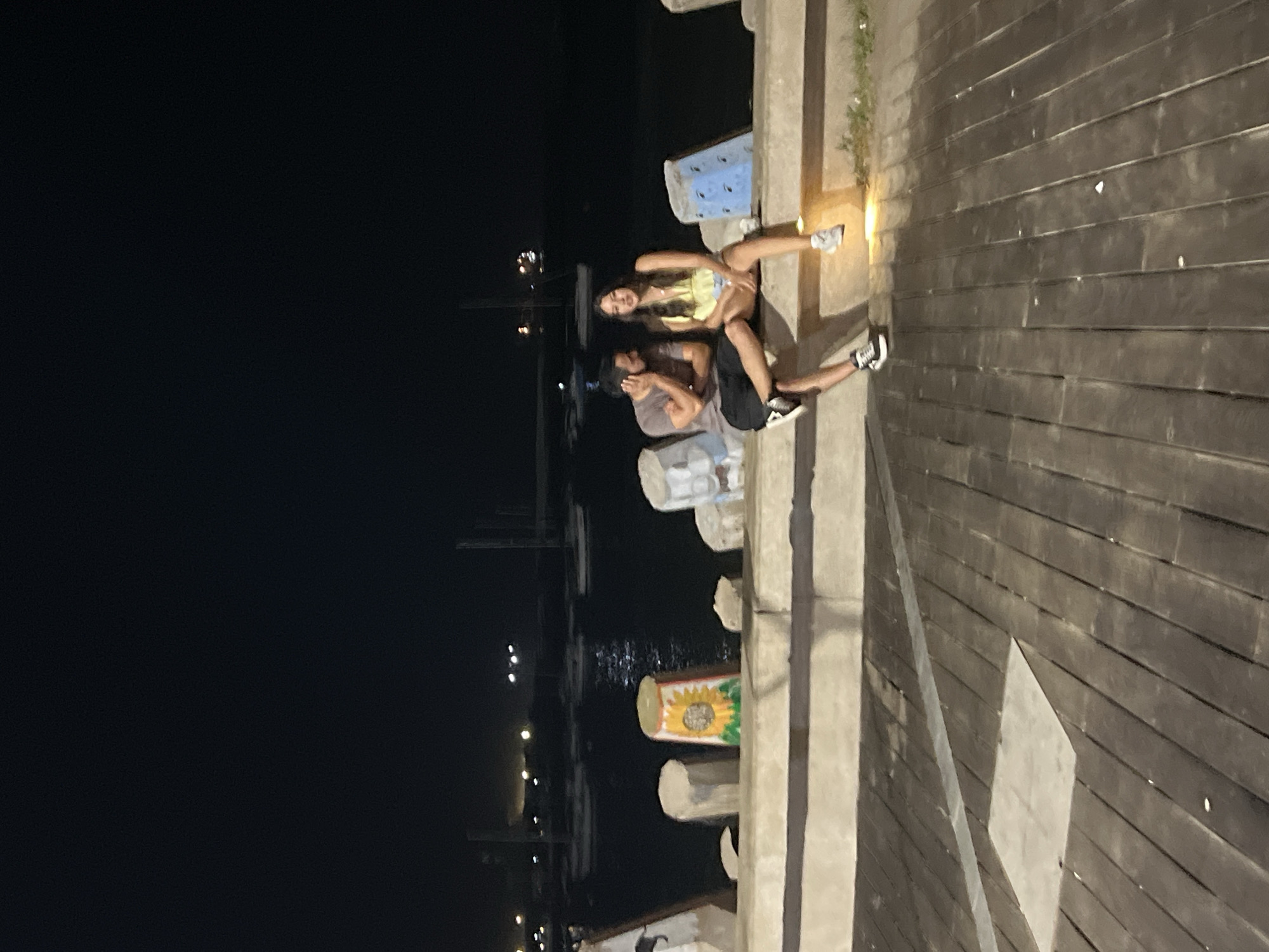 Two people are sitting on a pier or dock at night. A woman is seated above a man, with her legs extended. The man appears to be looking to his left. The dock is constructed of wood planks. There are concrete posts along the edge of the pier. In the background, lights are visible across the water, indicating an urban setting. The city is Arica, Chile.