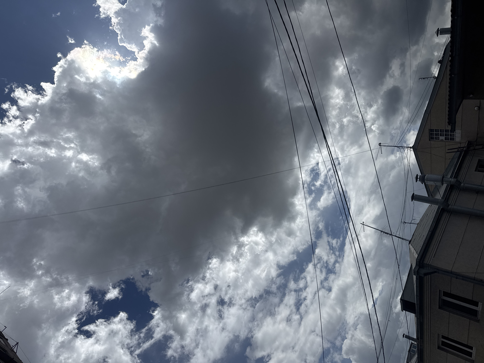 The image, taken from a low angle in Yerevan, Armenia, shows a sky filled with mostly gray and white cumulus clouds, with patches of blue sky visible. Electrical wires cut diagonally across the frame. The corner of a building with windows, pipes, and chimneys is visible in the upper right and lower left of the frame. The building appears to be a multi-story residential structure. The lighting suggests it is daytime. There are no people visible.