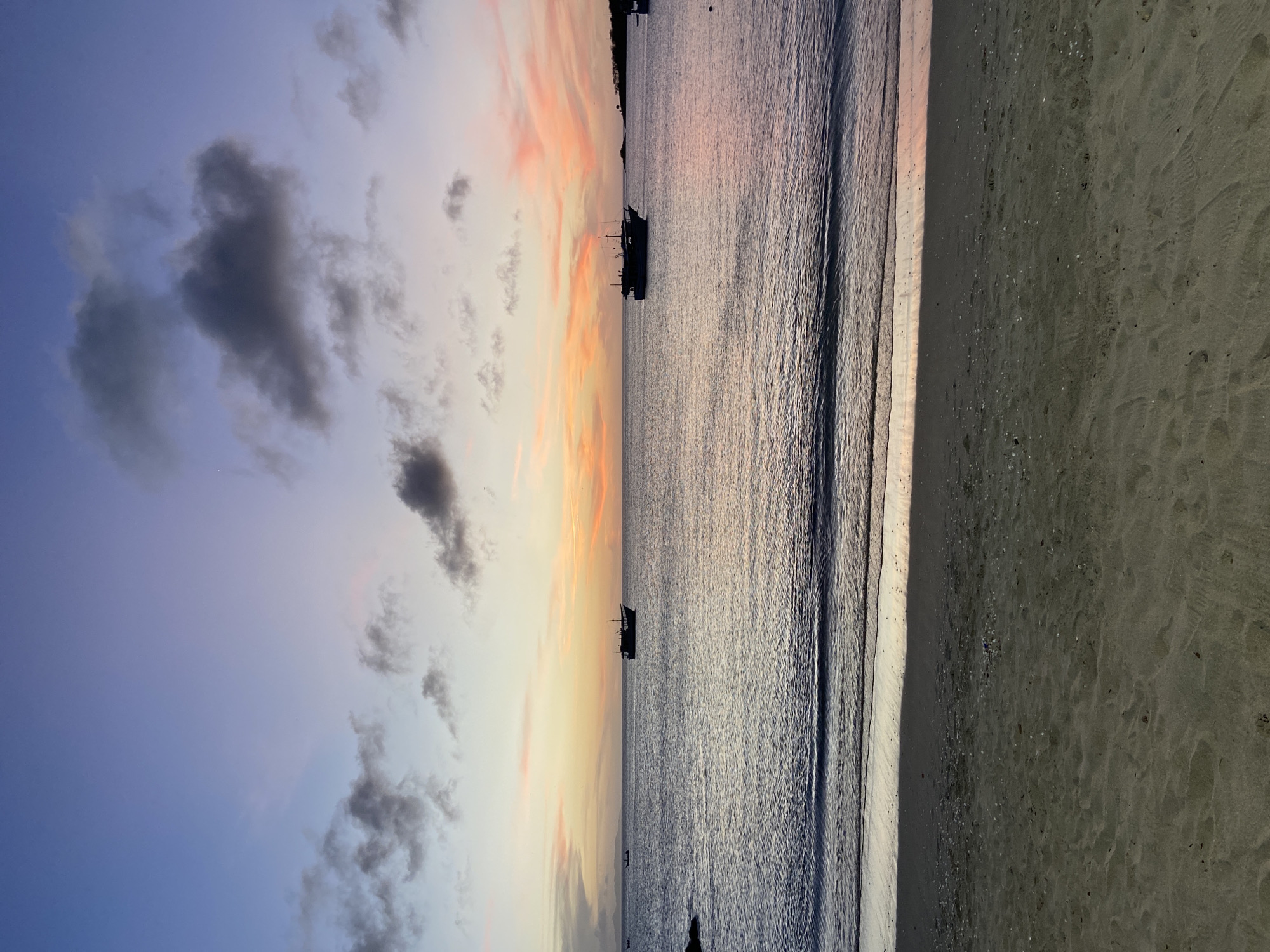 The image, correctly viewed horizontally, captures a tranquil sunset or sunrise over a beach in Juan Griego, Venezuela. The sky occupies the upper portion of the frame, showcasing a dramatic color gradient. On the left, it transitions from a deep indigo-blue to softer purples and then to vibrant oranges and pinks closer to the horizon on the right. Scattered dark grey clouds are visible across the sky, particularly in the bluer sections.
The calm sea fills the middle ground, its surface gently rippled and reflecting the soft hues of the sky, giving it a luminous, metallic appearance. Several small boats are silhouetted against the bright horizon line, suggesting local fishing or leisure vessels. One boat is more clearly visible in the mid-distance on the water.
The sandy beach makes up the rightmost section of the image. The sand is dark, possibly wet from the receding tide, and displays subtle textures and scattered small shells or debris along the water's edge. A distinct line marks where the gentle waves meet the shore.
The overall scene is devoid of visible human activity, contributing to a sense of peaceful natural beauty. The low, warm light indicates either dawn or dusk, creating a serene and contemplative atmosphere. There is no visible text in the image.