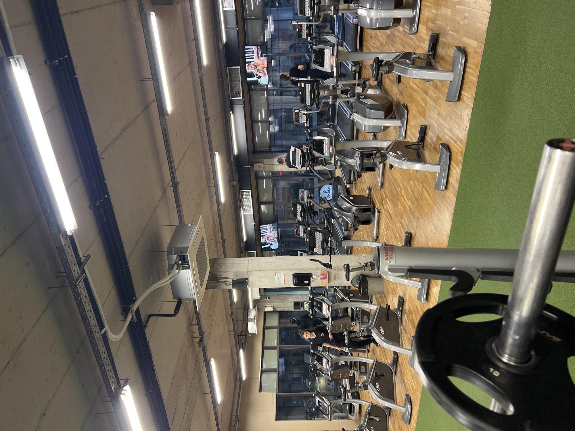 The image shows the interior of a gym, likely in Sofia, Bulgaria. Rows of elliptical machines and treadmills are arranged on a wooden floor, with a green artificial turf area visible. Several people are using the equipment, and a television screen displays content in the background. Overhead, fluorescent lights illuminate the space, and there's exposed ductwork and conduit. In the foreground, a weight plate is on a weight-lifting machine.