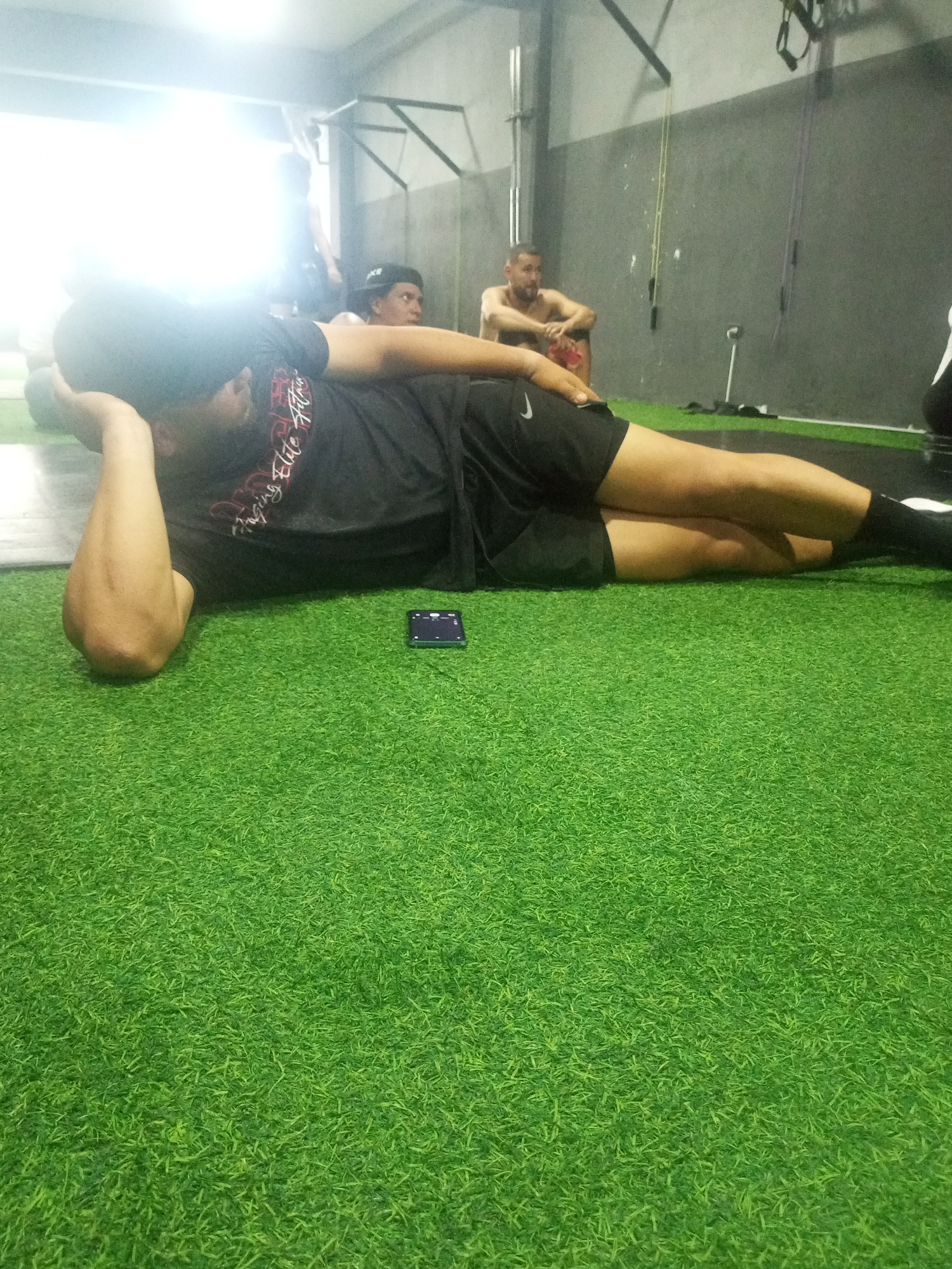 In a gym setting in Juan Griego, Venezuela, a man is lying on his side on a green turf floor, resting his head on his arm. He is wearing a black t-shirt and black shorts. To his left, another man sits on the floor, looking to the right. In the background, a third man is visible, leaning against a wall. The gym appears to be equipped with exercise apparatus, including resistance bands hanging from the ceiling. A small black object, possibly a timer, is placed on the turf near the man who is lying down. The lighting suggests indoor illumination, with a bright light source visible in the upper left corner of the image.